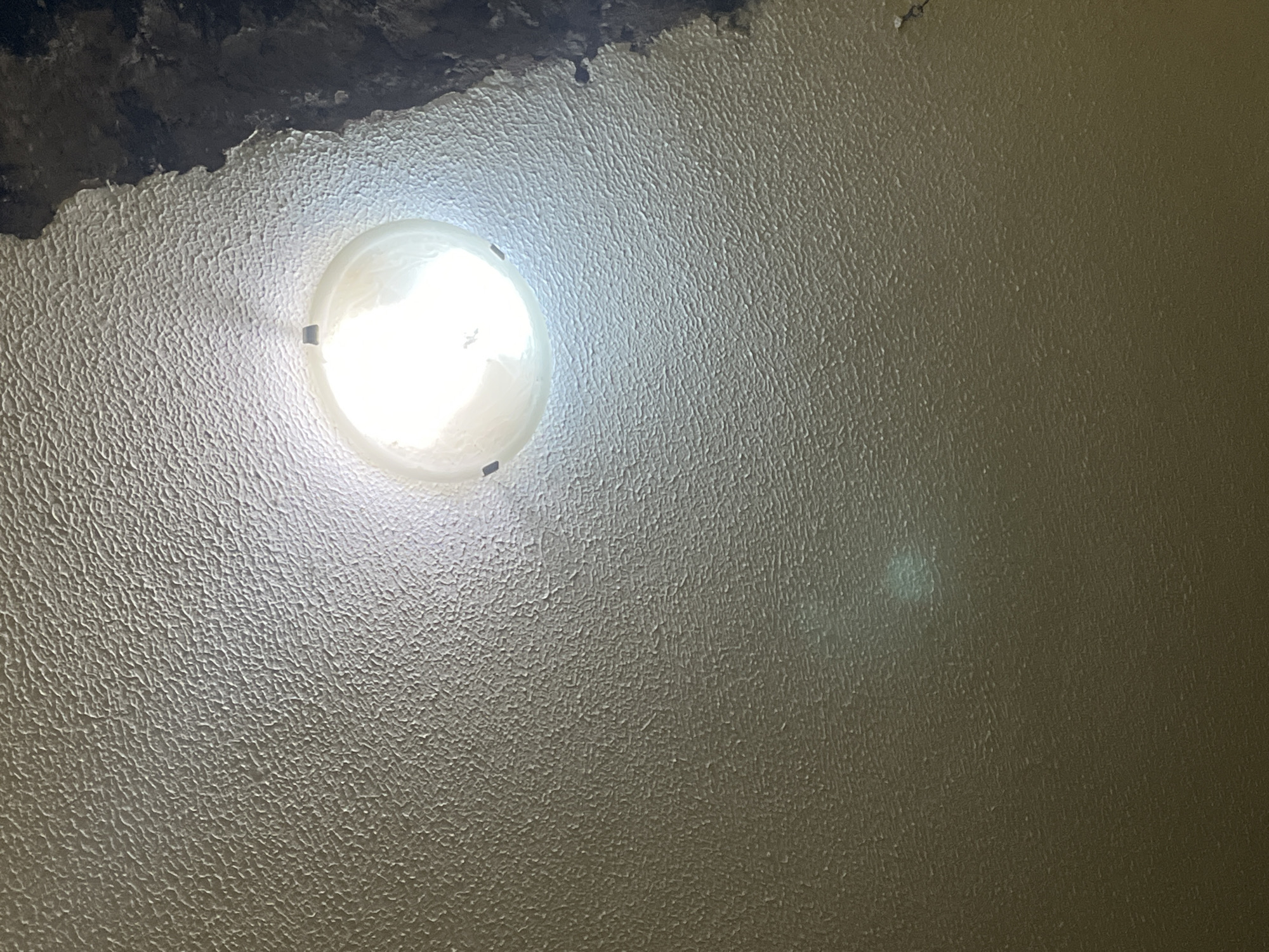 The image shows a close-up, upward-facing view of a textured ceiling with a light fixture. The ceiling has a rough, popcorn-like texture and is illuminated by a bright light emanating from a translucent dome-shaped fixture. The fixture appears to be circular or oval and is attached to the ceiling. There is a dark, irregular area in the upper right corner of the image, suggesting a hole or damage to the ceiling. The overall lighting is uneven, with the area around the light fixture being significantly brighter than the rest of the ceiling. There are no people or any other objects visible in the scene. The image does not provide any information that could be used to determine its location in Santa Ana, Venezuela.