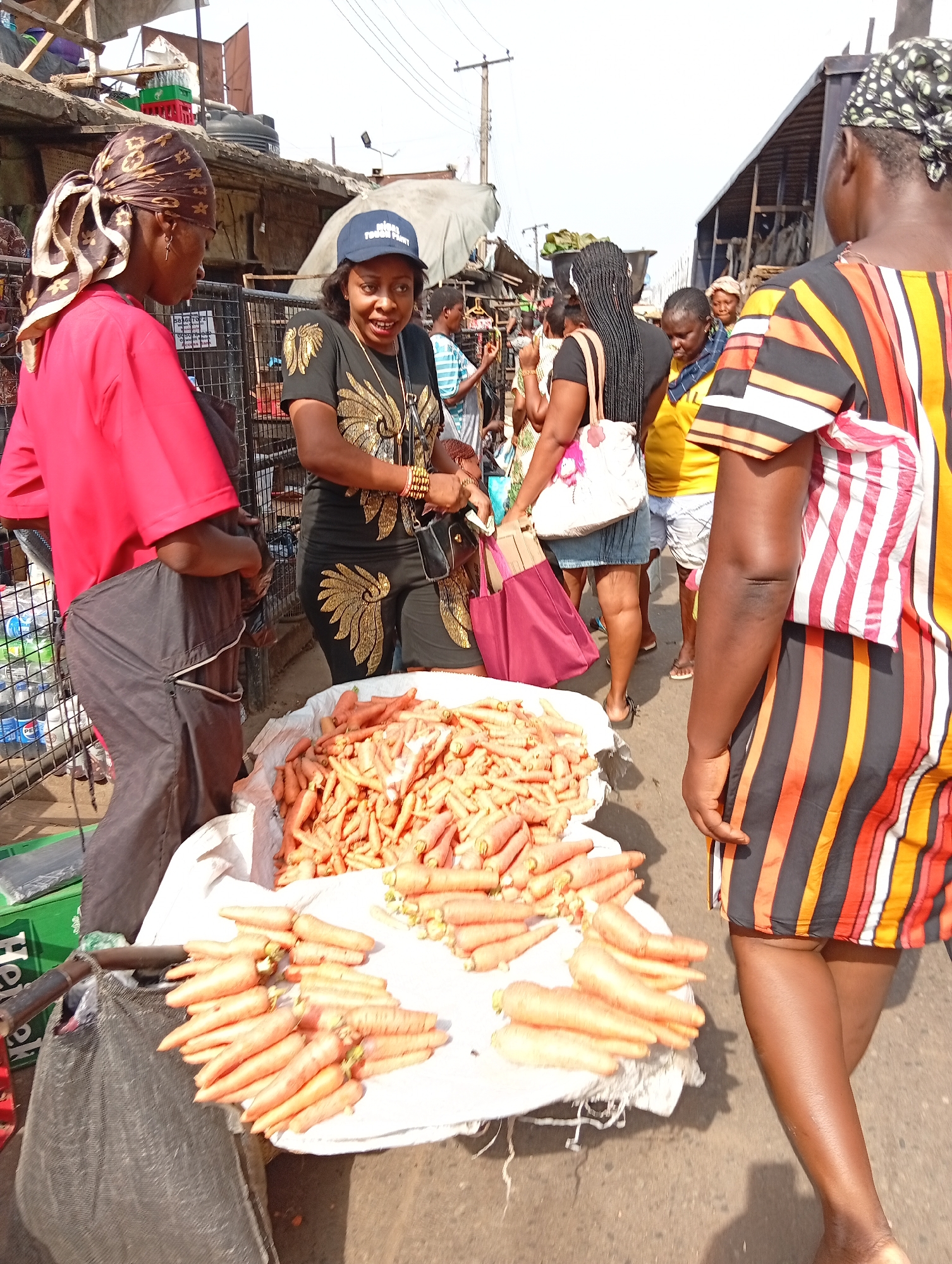 The image depicts a bustling market scene in Nkpor, Nigeria. A large pile of carrots sits on a white cloth atop what appears to be a low table or cart. Three women are prominently featured: one in a red shirt and dark pants, standing beside the carrots; another wearing a black shirt with gold embellishments and a blue baseball cap, seemingly interacting with the vendor; and a third in a striped orange, black, and white dress. The woman in the black shirt seems to be paying for the carrots.
Other people are visible in the background, including a woman in a yellow shirt and denim skirt. There are market stalls and buildings visible in the background, indicating a crowded commercial area. Crates of water bottles are visible next to the carrot stall. The image suggests a vibrant market setting with active buying and selling.