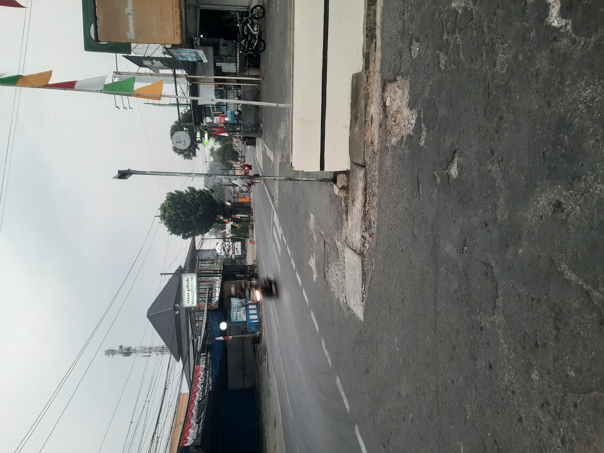 A street scene in Purwanegara, Indonesia, shows a road with a pedestrian crossing in the foreground. The road is paved and shows signs of wear, with cracks and patches. On the right side of the road, there is a white wall with a black stripe. Behind the wall are buildings and shops. Several flags, including Indonesian flags, are displayed. A motorcycle is visible on the road, blurred due to motion. A person is walking on the sidewalk on the left side of the road. In the background, there is a tall telecommunications tower. The sky is overcast. The general environment appears to be a commercial area on a street with buildings and traffic infrastructure.