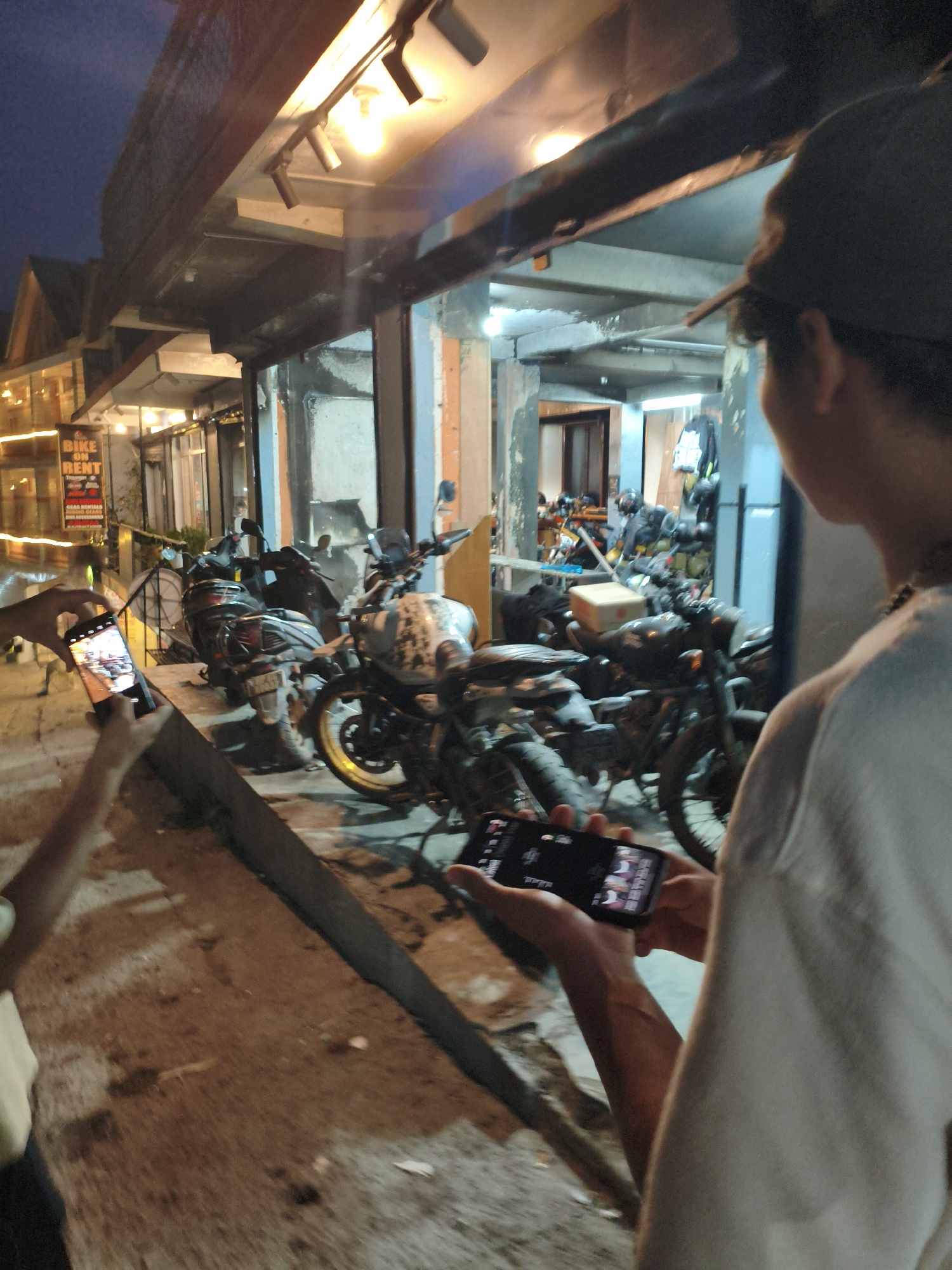A group of people are gathered outside a building that appears to be a motorcycle rental shop in Vashisht, India. Several motorcycles and scooters are parked on a raised concrete platform in front of the shop. The lighting suggests it is dusk or early evening. Two individuals in the foreground are holding smartphones, appearing to capture photos or videos of the motorcycles. The shop is illuminated by interior lights and overhead fixtures. One person is wearing a dark baseball cap.