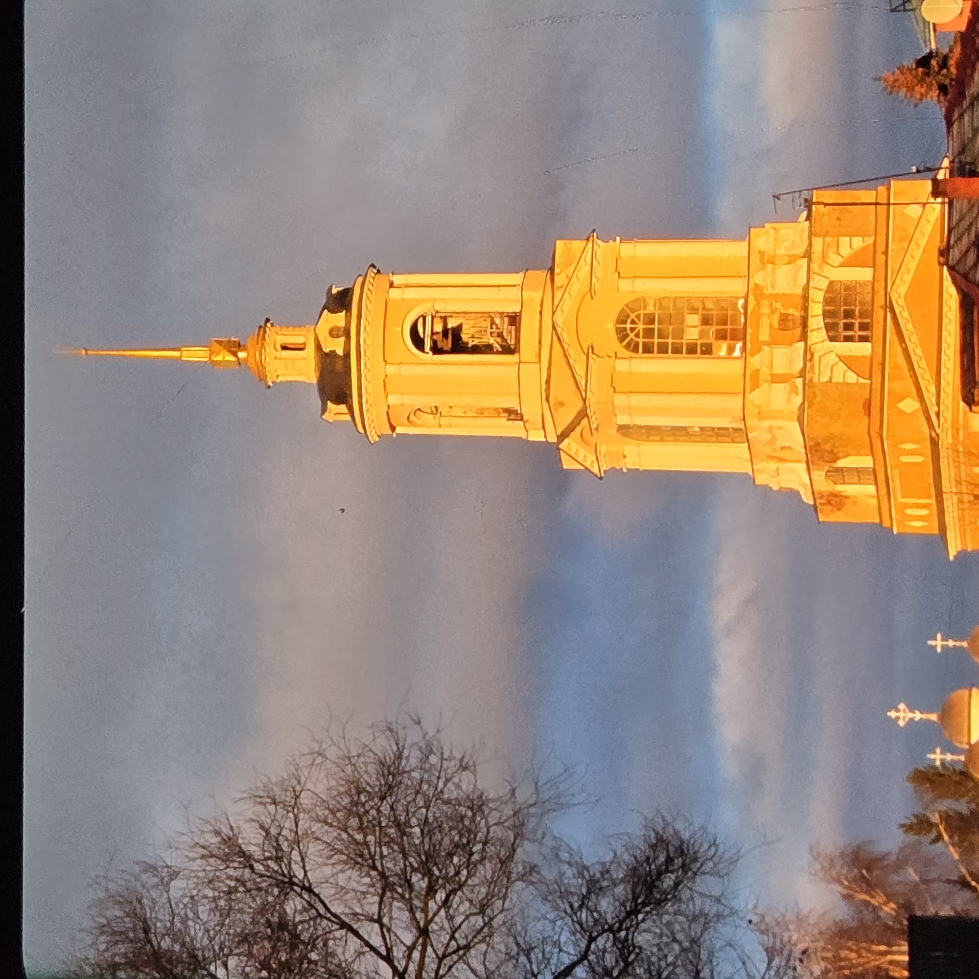 A golden-lit bell tower of a church stands prominently against a twilight sky in Vladimir, Russia. The tower, illuminated by the setting sun, has a slender spire topped with a cross. It features arched windows and decorative architectural elements. To the left, the bare branches of a tree are silhouetted against the sky, adding a stark contrast to the bright structure. In the lower part of the image, other church domes with crosses are visible, also catching the warm light. The sky is a gradient of blue and gray with hints of pink from the sunset, and some clouds are present.