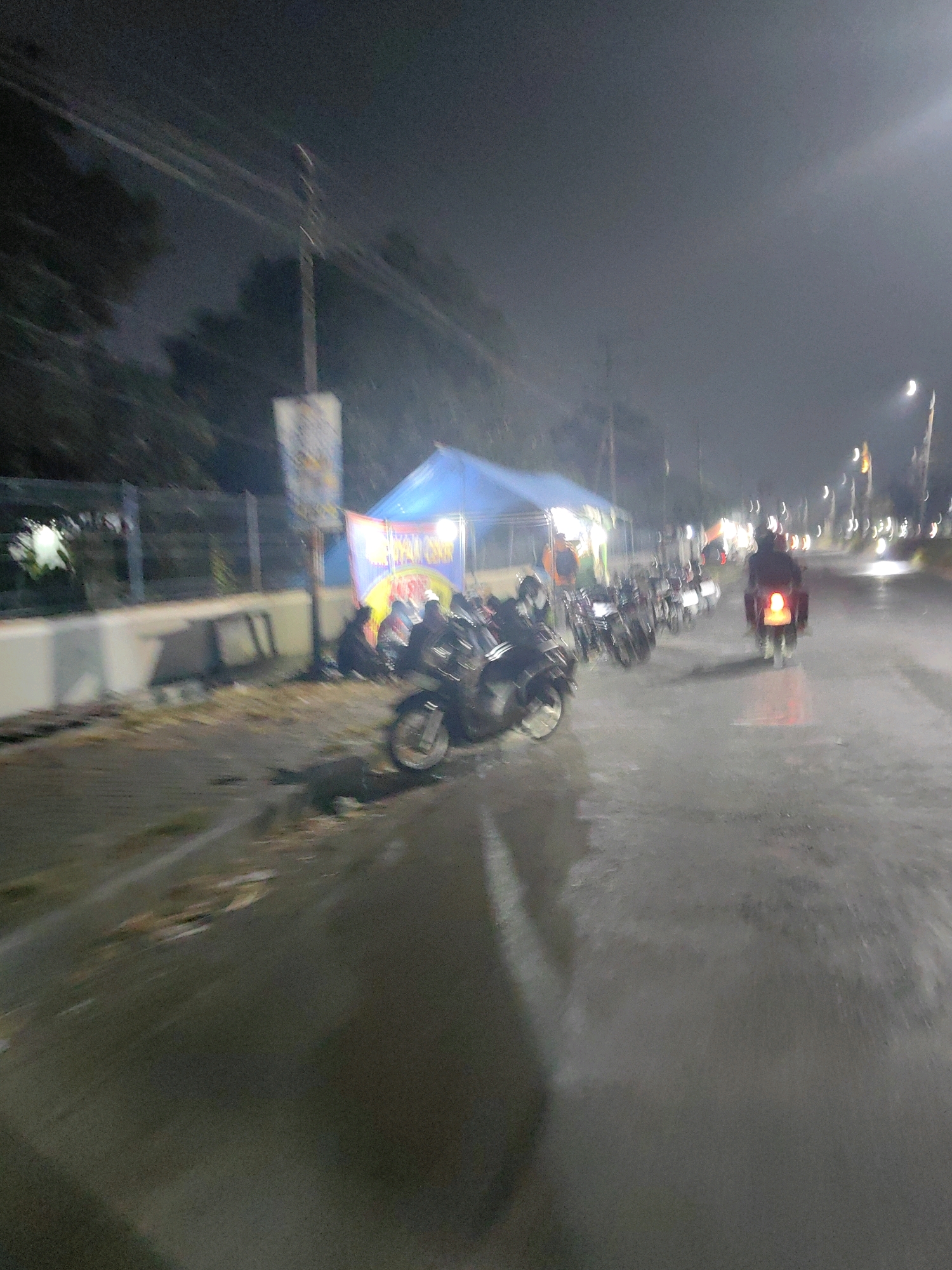 The image depicts a nighttime street scene in Keboansikep, Indonesia, likely near a roadside market or gathering. A blue tent is visible, possibly serving as a vendor stall or shelter. Several motorcycles are parked along the side of the road, and one motorcycle is in motion, its brake light illuminated. The road surface appears wet, suggesting recent rain or damp conditions. The overall lighting is dim, with artificial lights from the tent and street lamps creating reflections on the wet pavement. The scene is somewhat blurry, indicating movement or low light conditions. The presence of a pole with a banner and some individuals gathered near the tent suggest a community or commercial activity is taking place.