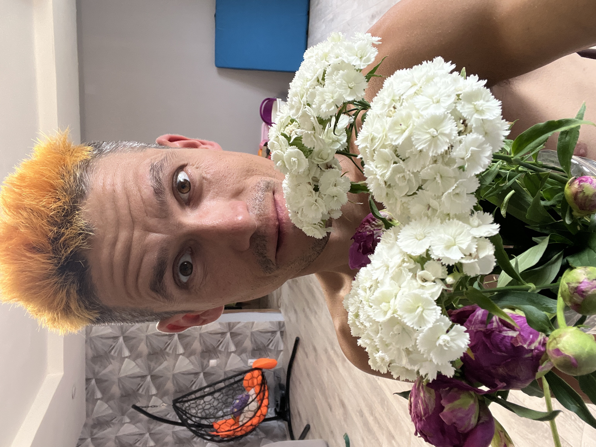 The image is a close-up, Dutch-angle shot of a person holding a bouquet of flowers. The person has short, spiky, orange-dyed hair and a surprised or concerned expression on their face, with wide eyes. They appear to be in an indoor setting, possibly a home, with a tiled wall and a patterned wall in the background. A blue object, possibly a mat or piece of furniture, is visible on the wall behind them.
The bouquet consists of white, fluffy flowers and several deep purple, closed flower buds, possibly peonies. The flowers are held in front of the person's face, partially obscuring it. The lighting suggests it is daytime, and there are no explicit indications of weather conditions.
The scene feels personal and candid, capturing a moment of interaction between the person and the flowers. The person's expression is the focal point, conveying a sense of surprise or being startled. The background details, like the patterned wallpaper, suggest a domestic environment. There is no visible text in the image.