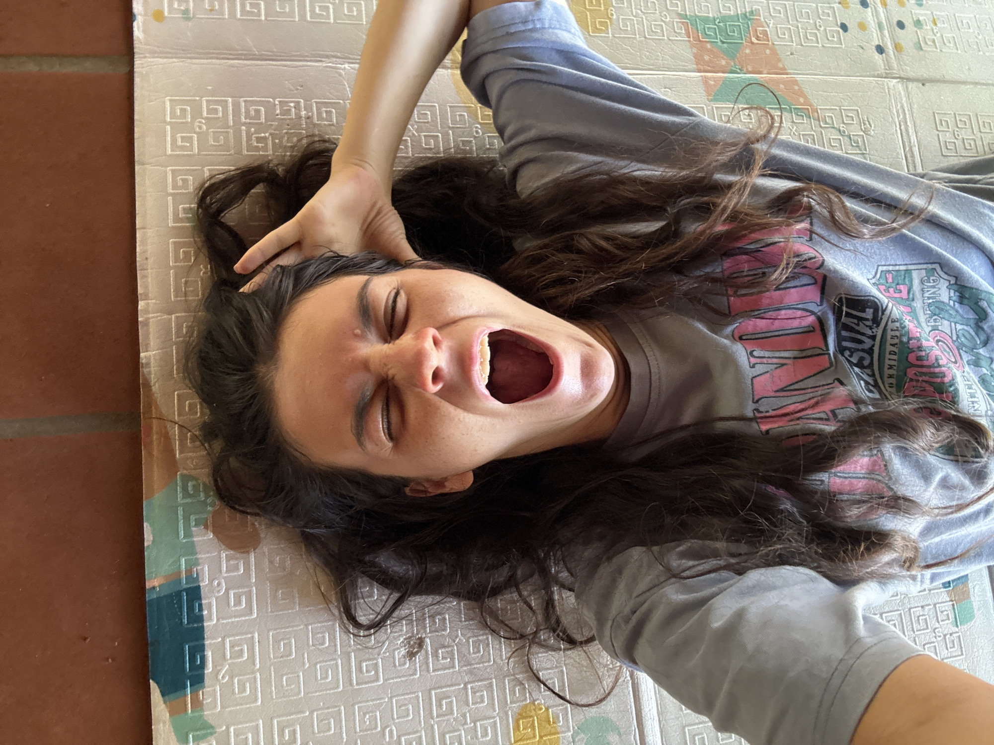 A young woman lies on a padded mat, her head turned to the left and mouth wide open, possibly yawning or shouting. Her hair is dark and wavy, spread around her head on the mat. She is wearing a grey t-shirt. Her left arm is raised and her hand is touching her hair. The mat appears to be composed of interlocking foam tiles with a decorative pattern. A portion of a tile floor is visible on the left edge of the frame. There is no evidence that indicates the location is in Mũi Né, Vietnam.