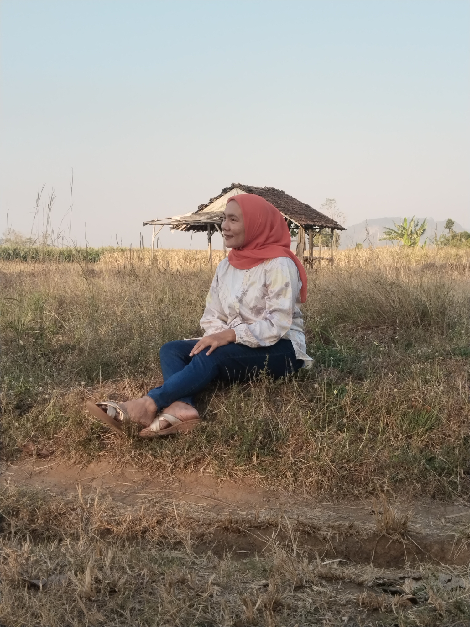 In the city of Kaliwungu, Indonesia, a woman wearing a peach-colored hijab, a long-sleeved patterned shirt, and blue jeans sits on dry grass in a field. She is wearing flat sandals and is looking to her right with a slight smile. Behind her, a small, rustic structure with a thatched roof sits in the background, surrounded by dry vegetation and some distant trees. The sky is clear and pale. The ground in the foreground shows some disturbed earth and patches of green and dry grass.