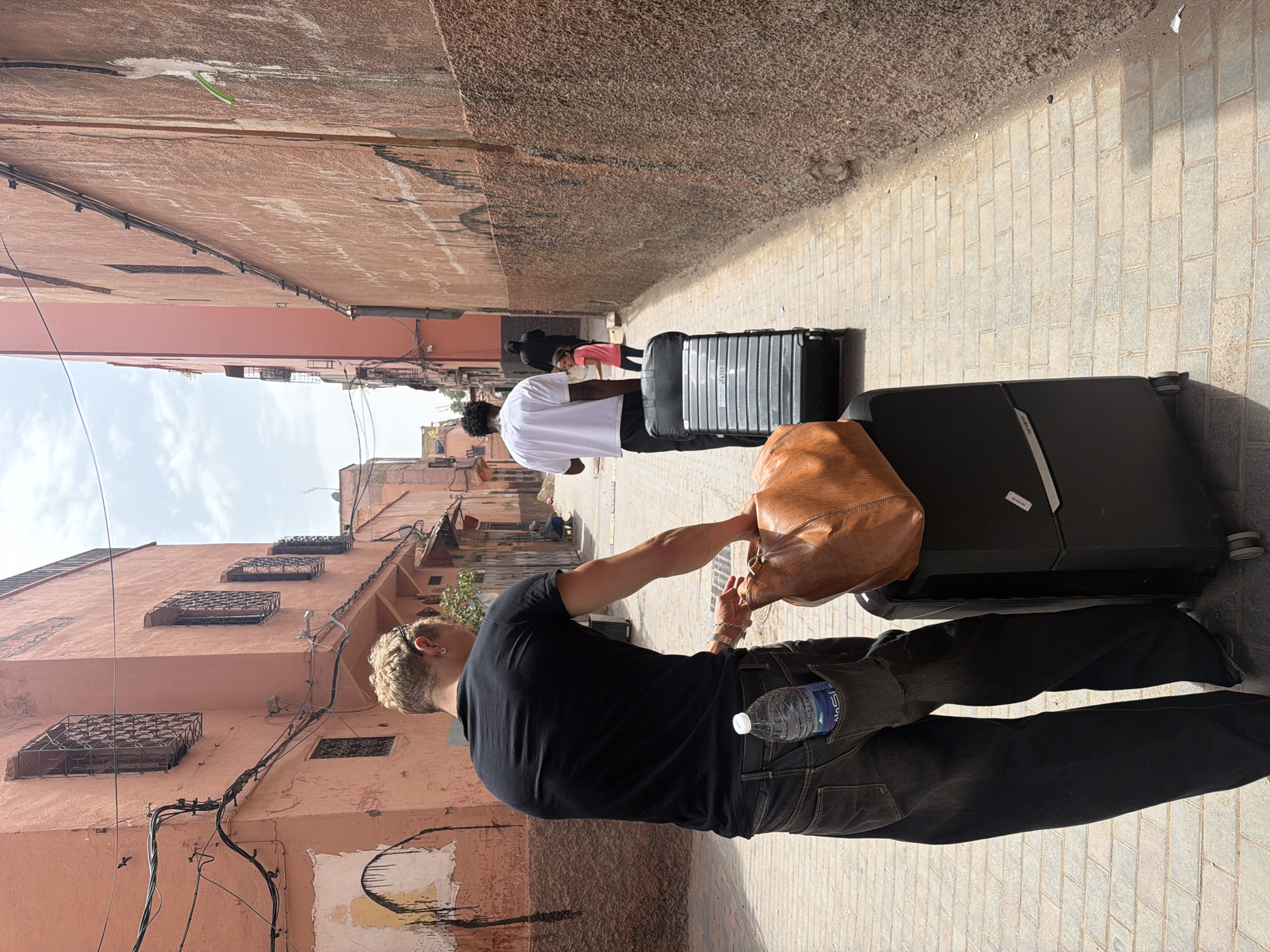 This image depicts a narrow alleyway in what appears to be a bustling medina, likely in Marrakesh, Morocco, given the architectural style. The buildings lining the alley are painted in warm, earthy tones, with exposed brick and weathered plaster. Ornate metalwork on windows and the presence of numerous electrical wires add to the character of the scene. The ground is paved with cobblestones.
The primary subjects are two individuals, seen from behind. The foreground figure is a man with short, blonde hair, wearing a black t-shirt and dark jeans. He is carrying a brown leather duffel bag over his shoulder and has a bottle of water tucked into his back pocket. He appears to be interacting with or observing the person ahead of him. Behind him, another person, wearing a white t-shirt, stands next to a large, black, wheeled suitcase. Further in the background, a smaller figure, possibly a child in a pink shirt, is visible next to another black suitcase. Two other figures are partially visible in the distance, one in dark clothing.
The activity appears to be travel or moving luggage through the alley. The lighting suggests it is daytime, likely mid-morning or afternoon, with bright sunlight casting shadows on the ground. The overall atmosphere is one of movement and daily life within a historic urban environment. The presence of luggage suggests travel.