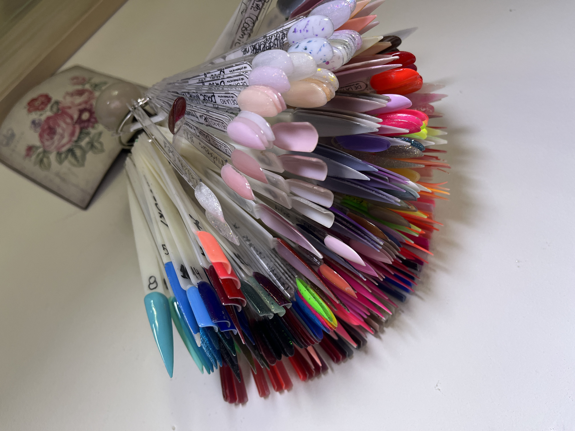The image shows a nail polish color sample display fanned out on a white surface. The display consists of numerous clear plastic sticks, each tipped with a different shade or design of nail polish. The colors range from light pastel pinks and purples at the top to vibrant reds, blues, and greens towards the bottom. Some samples have glitter or other decorative elements. On the left side, a decorative floral box is visible, resting against the fan of nail polish samples. The clear plastic sticks have product names and numbers printed on them, such as "DE LA RO". It's not possible to identify the location as Veliko Tarnovo, Bulgaria, based on the visual information provided.
