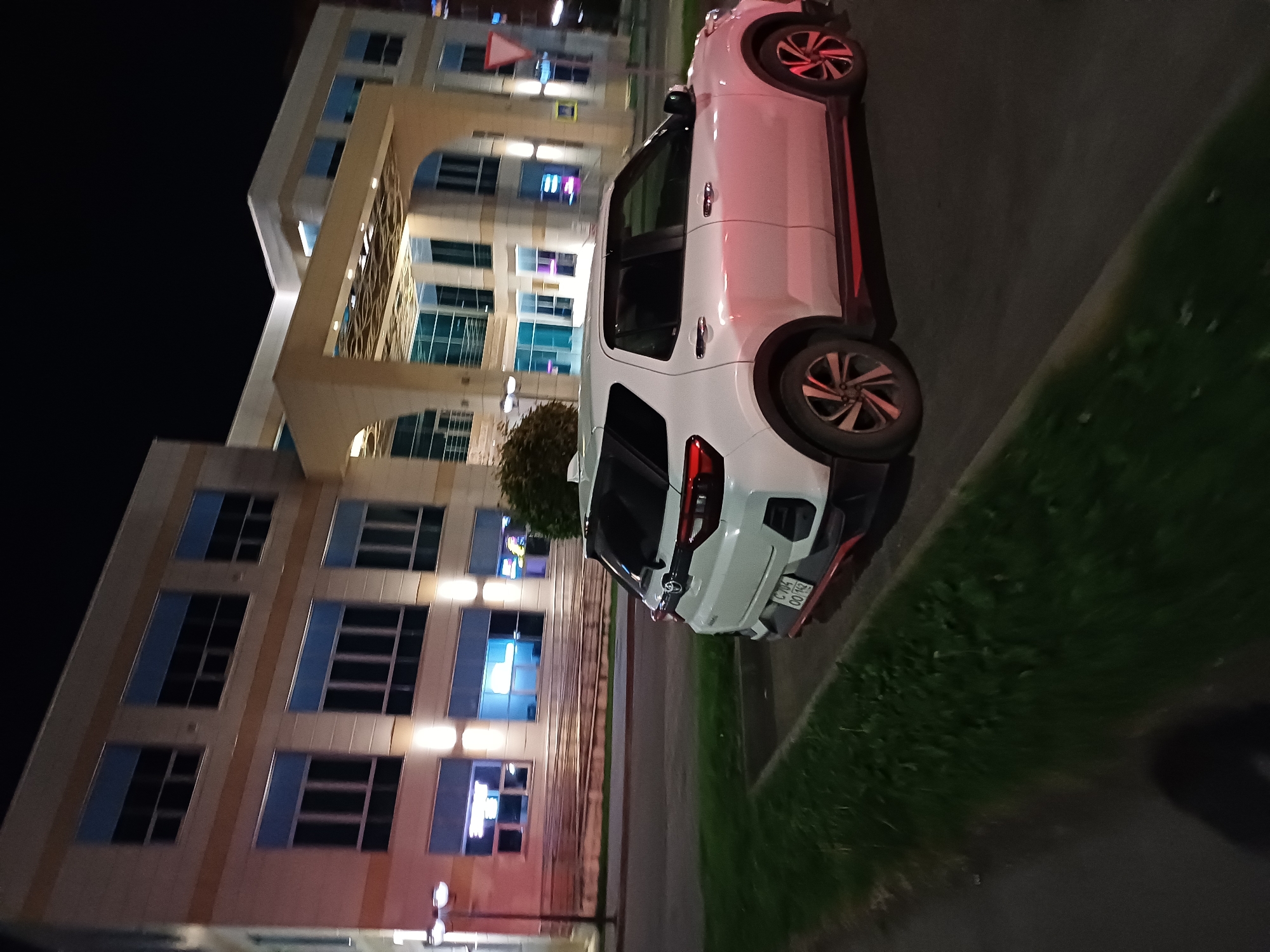 A white, four-door crossover SUV is parked in a dimly lit, paved lot at night. The car is facing right, and its rear is partially visible. The vehicle has a red accent strip along the bottom of its side and a red detail on the rear bumper. The car's license plate is visible and reads "C 704 00 1M3". Behind the car, a multi-story building with large, illuminated windows is visible. The building appears to be modern, with a beige facade and blue-tinted windows. Some of the windows show interior lighting, and neon signage is visible in a few. A well-maintained patch of green grass borders the parking lot on the left. Streetlights and the glow from the building illuminate the scene. The sky above is dark.