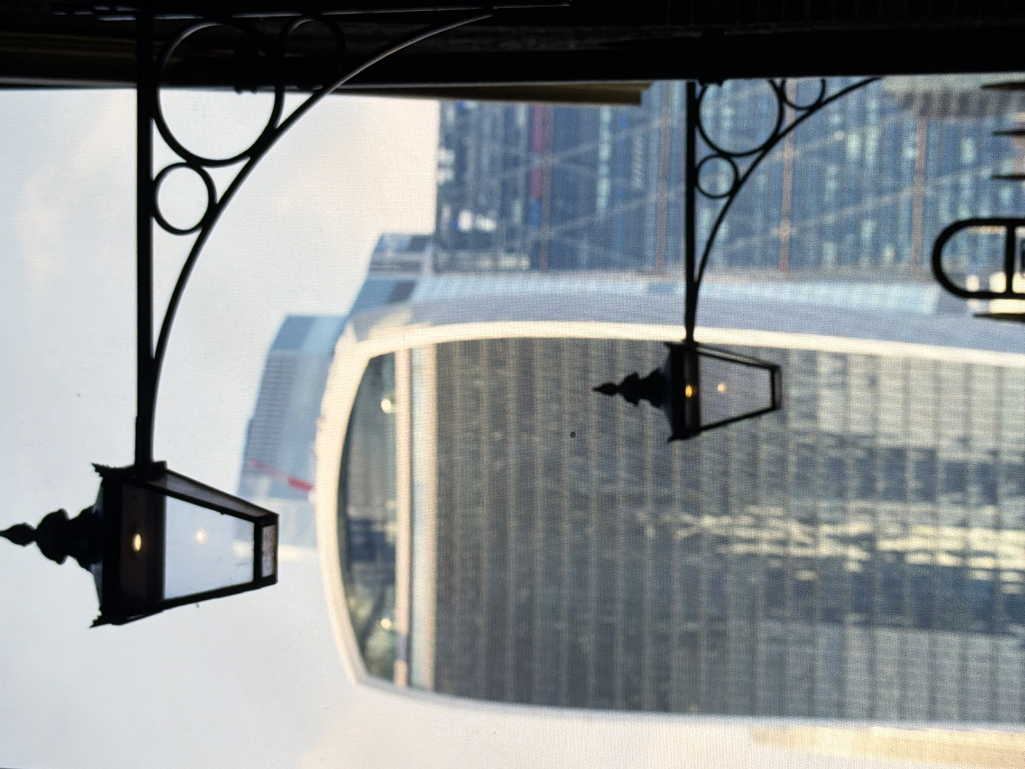 The image displays a detailed view of two dark, ornate street lamps silhouetted in the foreground against an out-of-focus urban background. The upper lamp, positioned in the top-left quadrant, is larger and features a pointed finial, clear glass panels, and an illuminated warm-toned light source within. Its support arm, extending right, includes decorative scrollwork and two dark circular elements. A second, smaller, similarly designed and illuminated lamp is visible mid-right with its own support structure. These lamps are affixed to a dark, vertical structure on the far right of the frame. Partially visible at the bottom center is another dark, arched metal object.
The background, seen through what appears to be a fine mesh or screen, reveals a modern city skyline under a bright, overcast sky. A prominent, large skyscraper occupies the center-left, distinguished by its convex, curved profile, wider at the top, and a facade of reflective glass with horizontal banding. To the right and further back, additional contemporary skyscrapers are visible, characterized by rectilinear forms and interior details or patterns showing through their glass exteriors, including hints of red and blue illumination.
The described scene is situated in Maidstone, United Kingdom.