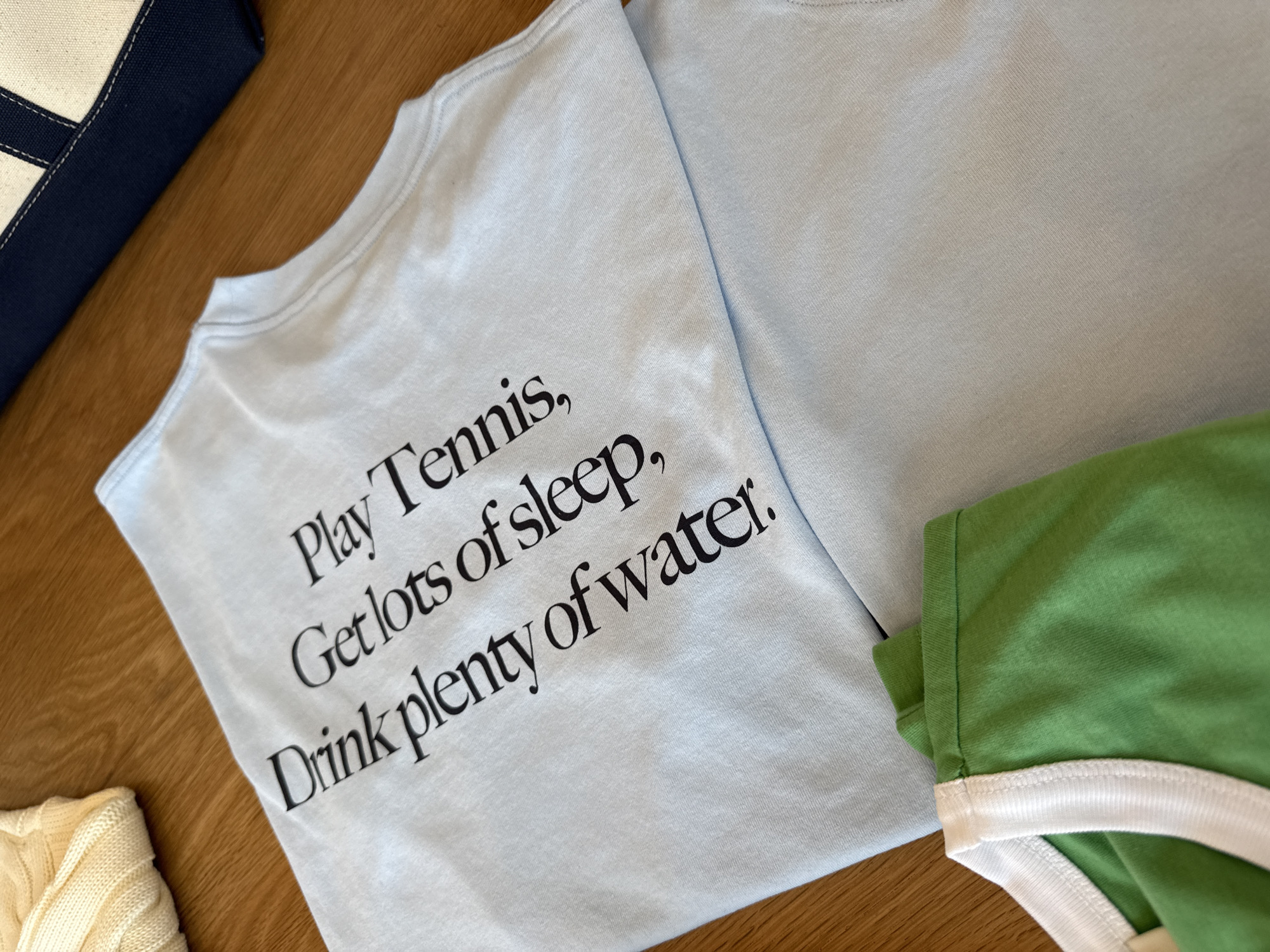 The image shows a light blue t-shirt with the text "Play Tennis, Get lots of sleep, Drink plenty of water." printed on it in black. A second light blue t-shirt is partially visible next to the first. To the right of the first t-shirt is a green ringer t-shirt with a white neckline. To the left is a blue and white bag, and a beige knit sweater. The image was taken indoors on a wooden surface. There is no evidence that the image was taken in West Hollywood, United States of America.
