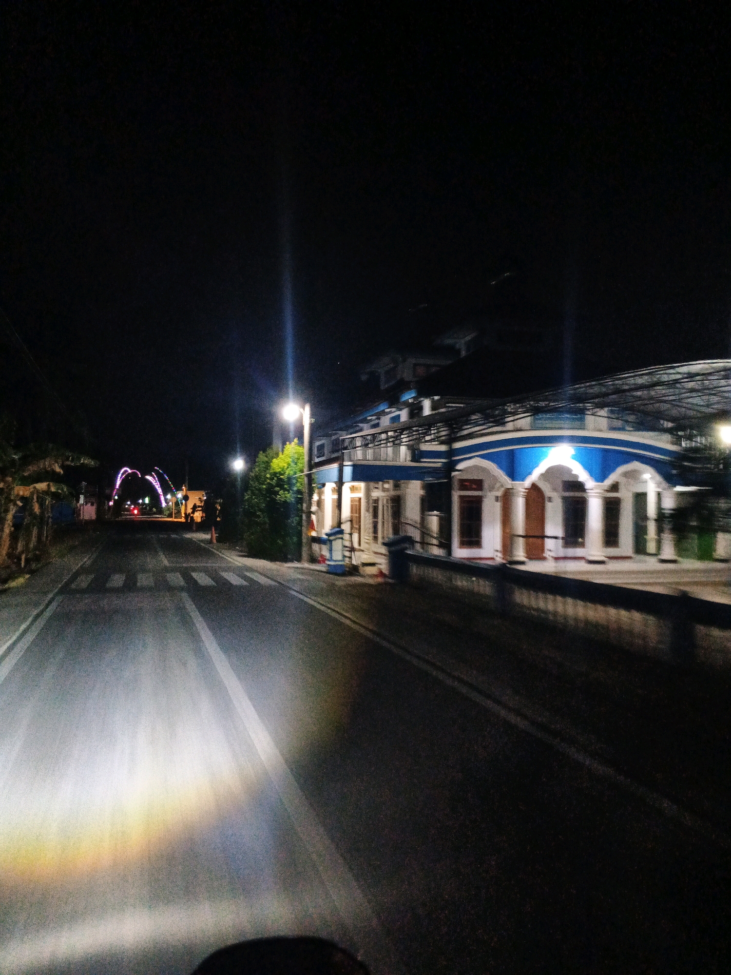 The image depicts a street scene at night in Sirau, Indonesia. The perspective appears to be from a moving vehicle, as indicated by the blurred motion of the road and the headlights illuminating the foreground.
On the right side of the street, there is a two-story building with a distinctive blue and white architectural design. The building features arched windows and pillars, and a bright light is visible from within. Electrical wires are strung across the upper portion of the building and extend into the darkness. A lamppost with a glowing light stands in front of the building. Next to the building, there are some blue trash receptacles.
On the left side of the street, there are trees and vegetation. Further down the road, an illuminated archway with colorful lights is visible, spanning across the street. Some red lights, possibly from a vehicle, are present near the archway. There is a pedestrian crossing marked on the road. Two figures, appearing to be people, are standing near the trees on the left side of the street, though their features are indistinct due to the distance and low light. The overall environment suggests a semi-urban or coastal setting, given the presence of vegetation and the open expanse of the road. The night sky is dark, with no visible stars.