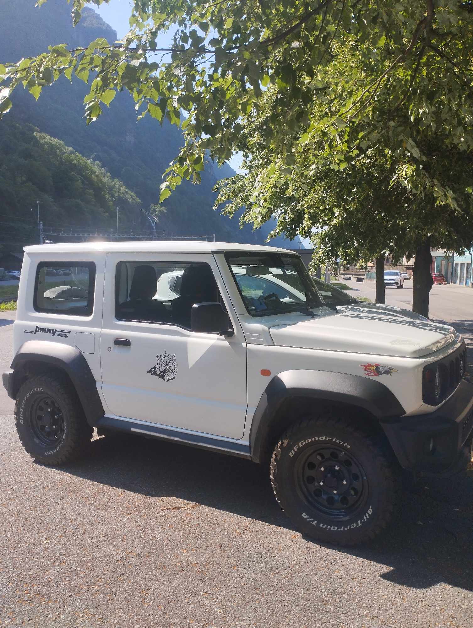 A white Suzuki Jimny is parked on a paved surface. The vehicle is a two-door SUV with black fender flares and black wheels. The car has a "Jimny" decal on the driver's side rear quarter panel and a compass decal on the same panel. There is also a decal of an eagle on the front fender. The car is parked under a tree with green leaves, and mountains are visible in the background. The sky is blue and clear. The scene appears to be in a mountainous area, possibly a town or village, with buildings and a road visible in the distance.