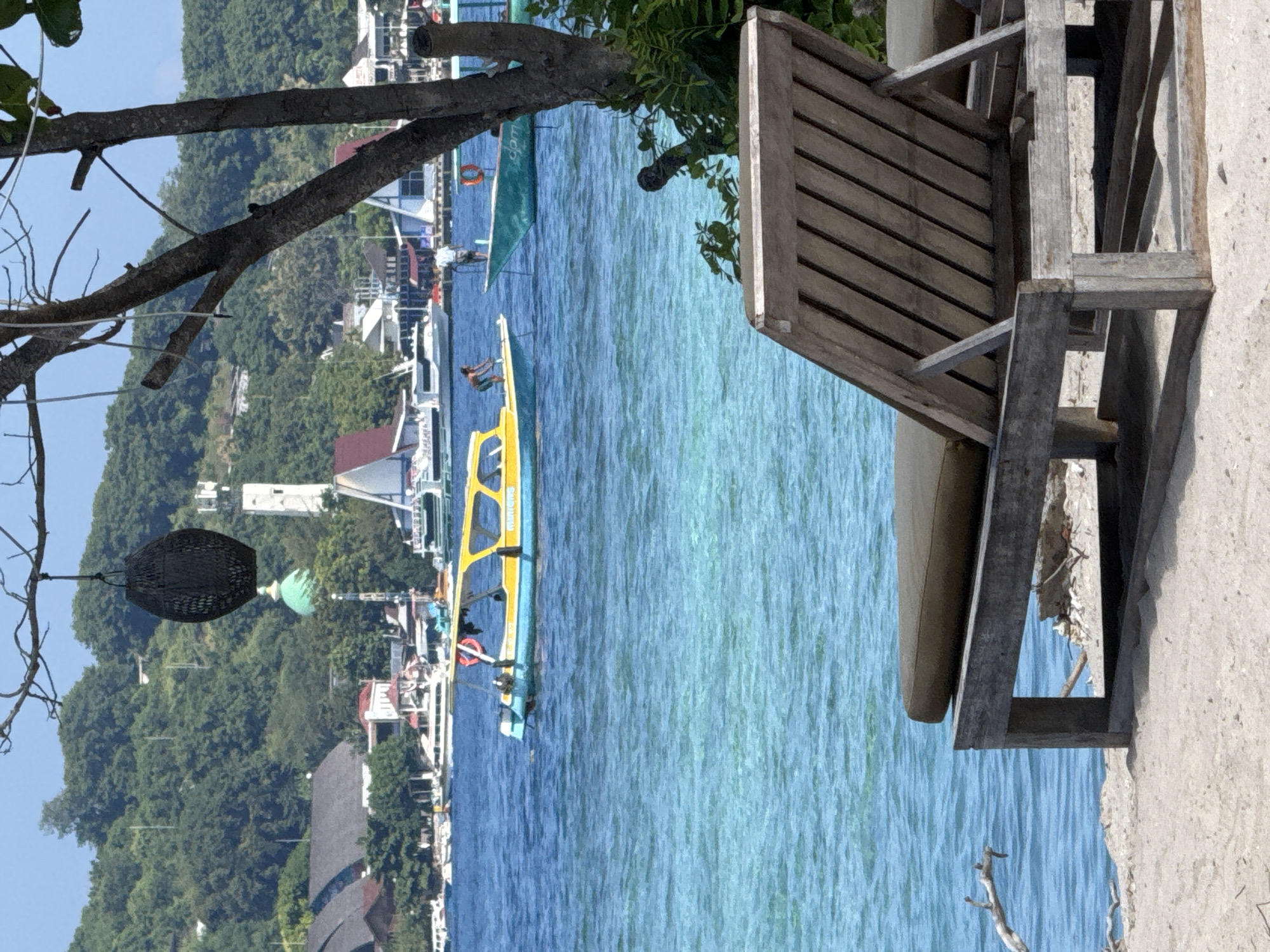 A vertically oriented, medium shot captures a coastal scene with a partially visible wooden chair in the foreground, obscuring the background of calm, clear blue water. A yellow and blue passenger boat is visible mid-frame, along with other vessels anchored near a densely vegetated shoreline dotted with buildings. The scene suggests a tropical, possibly tourist-oriented area. The sky is clear, indicating good weather conditions. The presence of a hanging woven object from a tree branch adds a local or cultural element to the scene. The photograph shows Unknown in Indonesia.