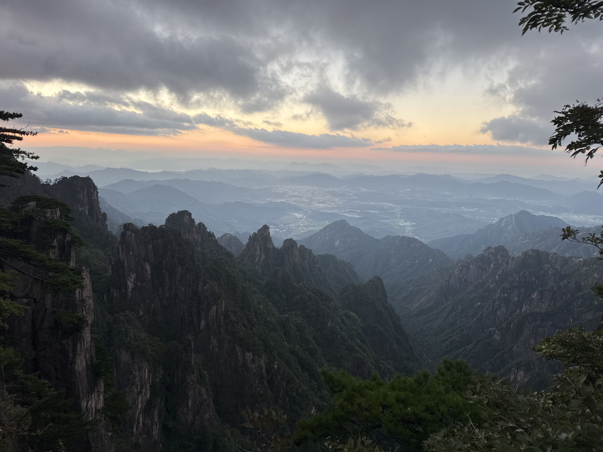 The image depicts a panoramic view of the Huangshan Mountains in Huangshan District, China, likely taken at dawn or dusk due to the soft, atmospheric lighting. Jagged, dark green, tree-covered peaks dominate the foreground and midground, with steep cliffs and deep valleys. In the far distance, a hazy landscape of rolling hills and a small settlement with faint lights is visible, blending into the horizon. The sky above is filled with dramatic, layered clouds, with hues of grey, white, and a soft orange and pink glow near the horizon, indicating the presence of the sun. The overall scene is one of natural grandeur and serene beauty.