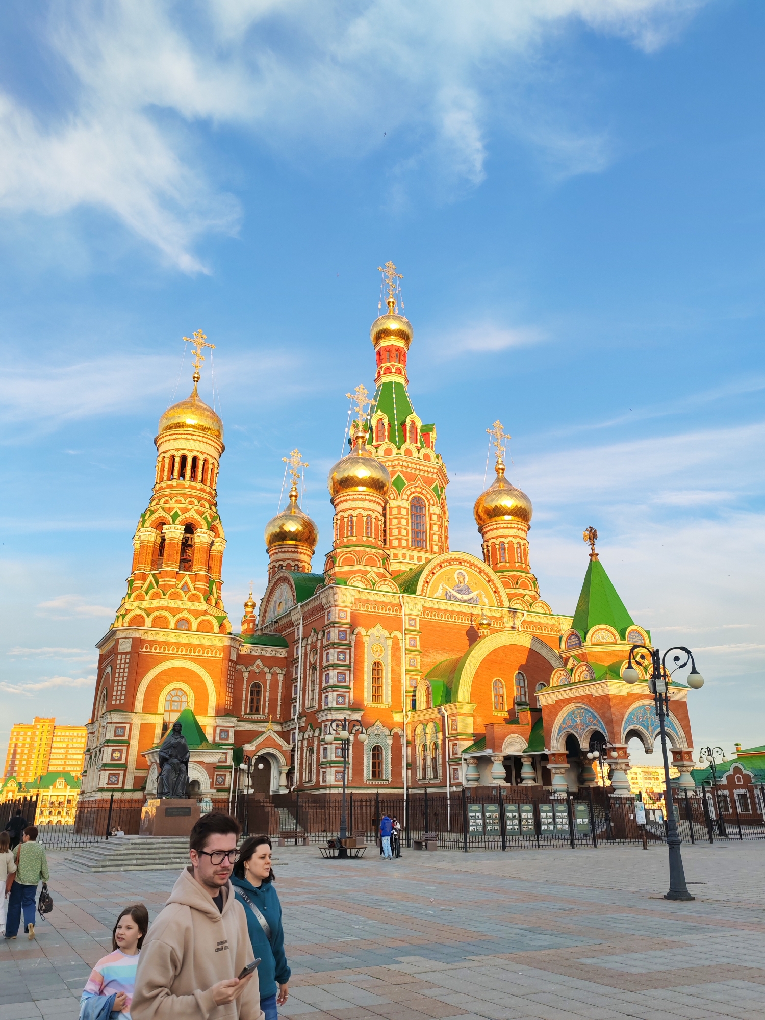 A wide shot captures the Church of the Nativity of Christ in Yoshkar-Ola, Russia, bathed in the warm glow of the setting sun. The vibrant orange and green facade of the onion-domed cathedral dominates the frame, its golden domes glinting against a bright blue sky dotted with wispy white clouds. In the foreground, a family walks across a patterned cobblestone plaza. A man in a beige hoodie and glasses looks at his phone, while a woman in a blue hoodie walks beside him. A young girl with a colorful striped shirt and a blue jacket in her arm follows them.
To the left of the church, a tall building complex stands in the distance, hinting at an urban setting. A statue of a figure in dark attire stands prominently on a pedestal in front of the church's entrance, overlooking the plaza. Black metal fences line the perimeter of the church grounds, with display boards visible behind them. A decorative lamppost with two globes stands to the right of the church, adding to the architectural detail of the scene. The overall atmosphere is serene and picturesque, with the late afternoon sun casting long shadows and highlighting the intricate details of the cathedral.