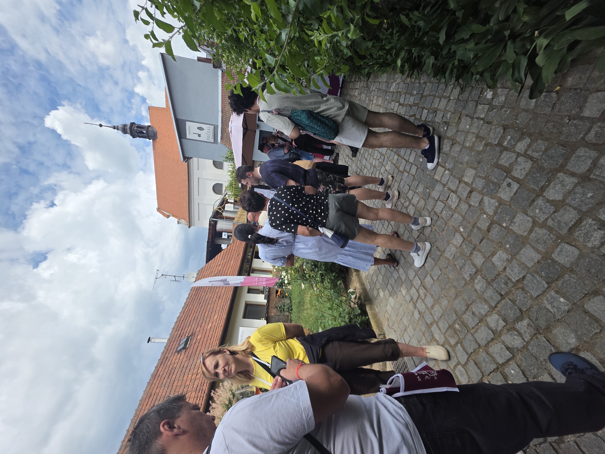 Here's a detailed description of the image:
**Scene Overview:**
The image shows a group of people standing on a cobblestone street. The photo appears to have been taken at street level, possibly by someone also standing in the group.
**People:**
Several individuals are visible, appearing to be of various ages. They are dressed in casual summer attire, including shorts, dresses, and t-shirts. At least one person is looking at a phone, while others seem to be engaged in conversation. A few people are standing in a line.
**Objects and Environment:**
* **Buildings:** Buildings with red-tiled roofs and light-colored walls are visible.
* **Sky:** The sky is partially cloudy.
* **Vegetation:** Trees or bushes are present on one side of the street.
* **Sign:** A sign is visible on one of the buildings.
* **Cobblestone Street:** The street is paved with grey cobblestones.
* **Tower:** In the background there is a building with a tower on top.
**Actions and Interactions:**
The people are standing and talking. They are looking at each other. Some of them look to be waiting in line.
