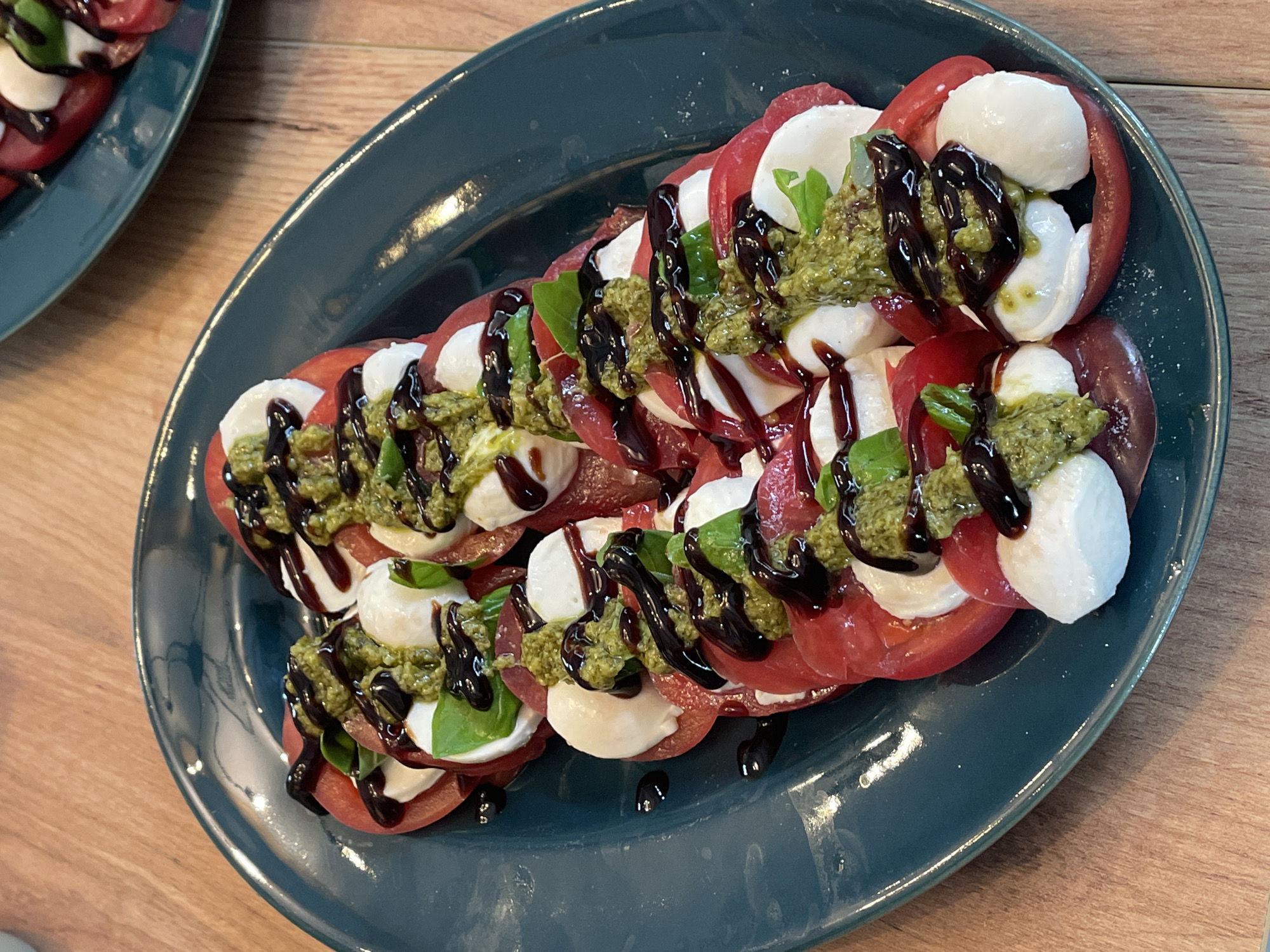 An oval, teal plate is filled with a Caprese salad, arranged in a fanned-out pattern. The salad consists of sliced red tomatoes and fresh mozzarella balls, layered and topped with basil leaves, a vibrant green pesto, and a dark balsamic glaze. A second, similar plate of salad is partially visible in the upper right background. The plates rest on a light brown wooden table.