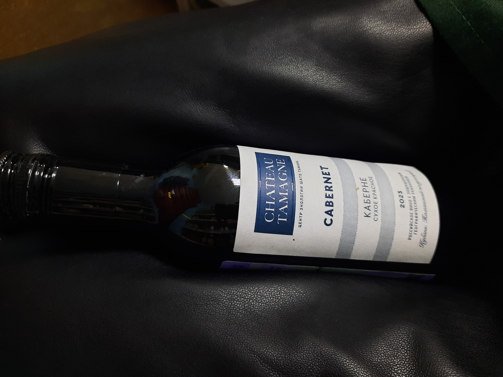 A bottle of Chateau Tamagne Cabernet red wine, vintage 2023, lies on a dark surface that appears to be leather. The label is white with blue text and vertical stripes. The text on the label is predominantly in Cyrillic. The wine is from Kuban, Tamansky region of Russia. The lighting is subdued and the bottle is angled slightly towards the viewer.