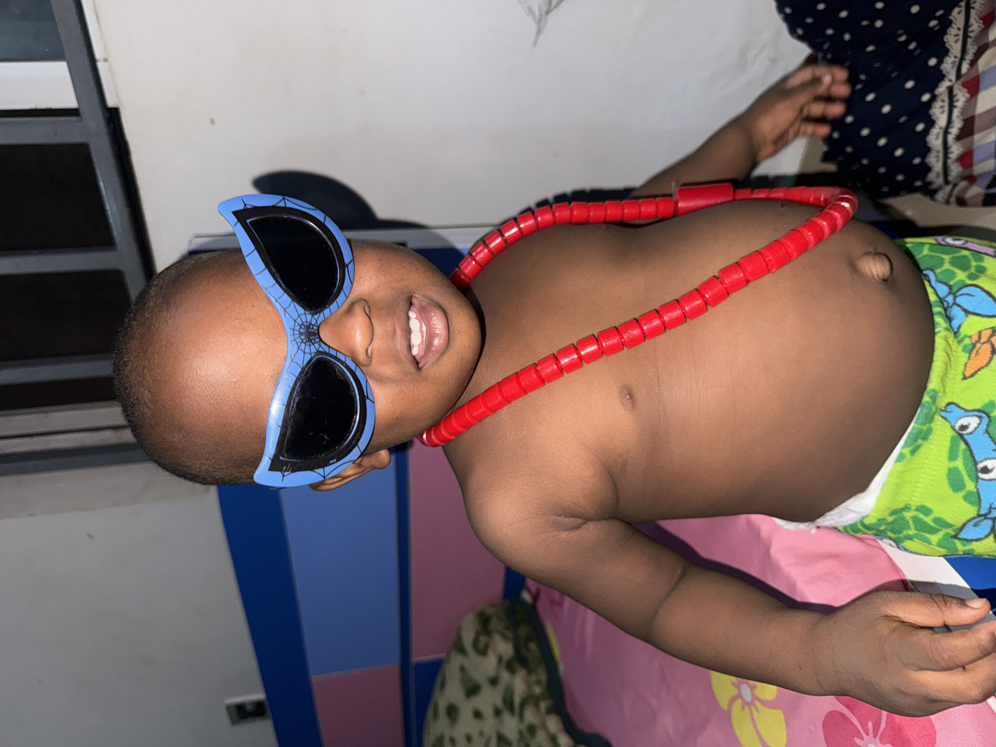 A shirtless child lies on a pink and green patterned surface, wearing blue spiderweb-patterned sunglasses and a red beaded necklace. The child's skin is dark brown. The child is lying on a bed or similar surface, with part of a green patterned diaper visible around their waist. A blue and white panel is visible to the left. The location is unknown, there is no evidence of Sabon Tasha, Nigeria.