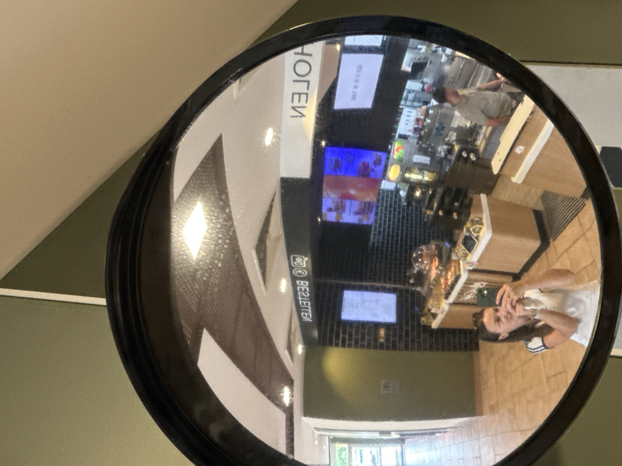 A round convex mirror displays the reflection of the interior of a building. Reflected in the mirror is part of a cafe or food court, showing a counter, display cases with food items, and people. One person, closer to the camera and presumably taking the picture, is holding a phone. The reflection suggests the room is brightly lit with various light fixtures. A sign in the reflection reads "HOTEL" (reversed) and "BESTELLEN" (reversed). The city is Ottersberg, Germany.