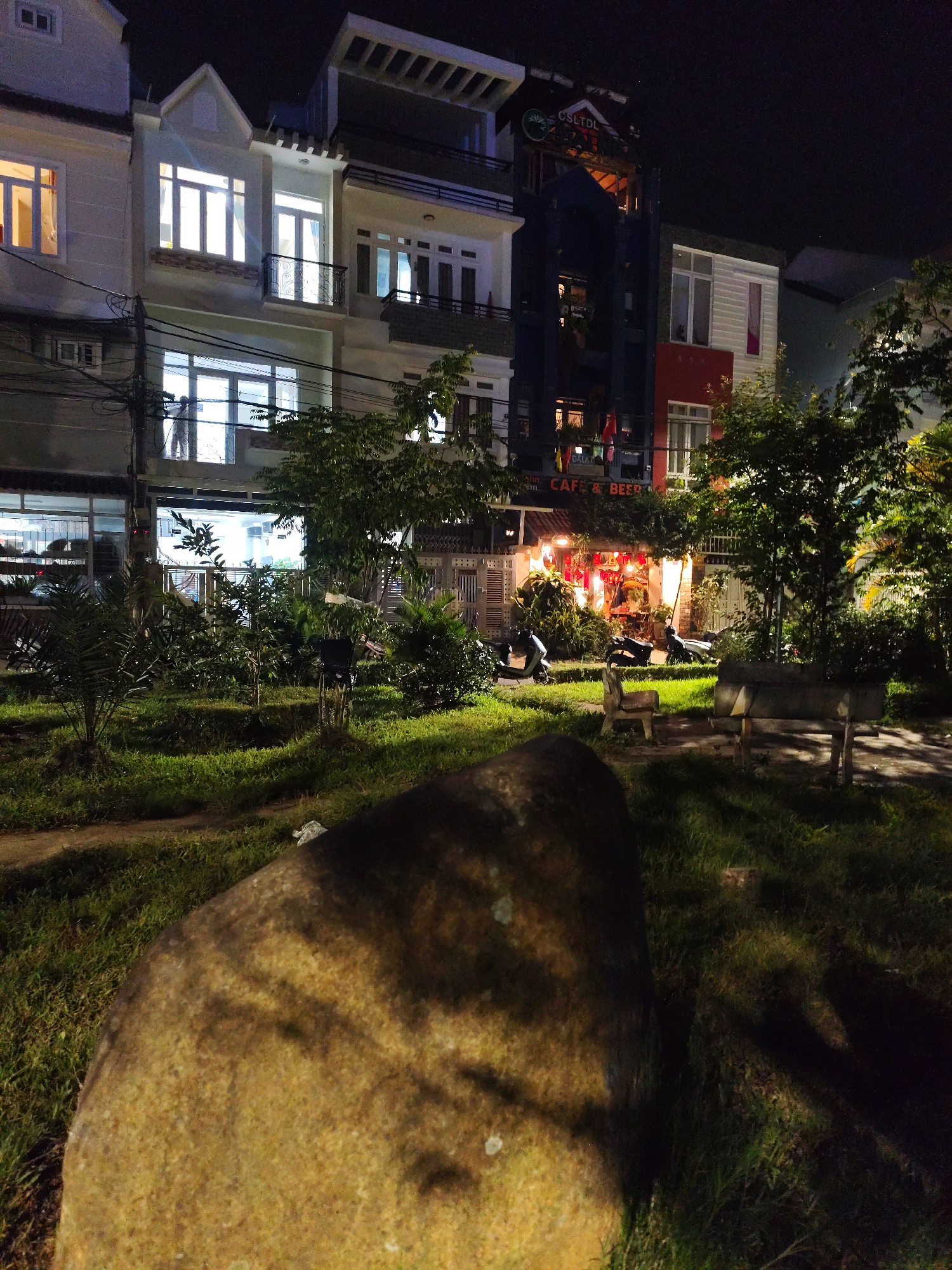 This nighttime photograph captures an urban scene in Da Lat, Vietnam, illuminated by artificial lights. In the immediate foreground, a large, textured boulder occupies the lower left corner. Beyond it, a grassy area with scattered small trees and bushes extends towards a row of buildings. Two simple concrete benches are visible in the midground, suggesting a public park or resting area.
The background features several multi-story buildings. On the left, a light-colored building has multiple windows radiating a warm, interior glow. Adjacent is another light-colored building with a small peaked gable; its brightly lit lower window reveals the faint silhouette of a person.
To the right, a dark blue building displays the partially visible text "CSLTDL" near its roofline and "HOSTEL" further down. Next to it stands a building with a red facade on its lower level and white siding above. At street level, below the dark blue building, a shopfront is brightly lit with warm lights and festive red lanterns. Visible text on its signage clearly reads "CAFE & BEER," with "DALAT" partially discernible below it. Several motorbikes are parked in front of these businesses. The sky is dark, indicating night, and the scene appears calm and quiet.