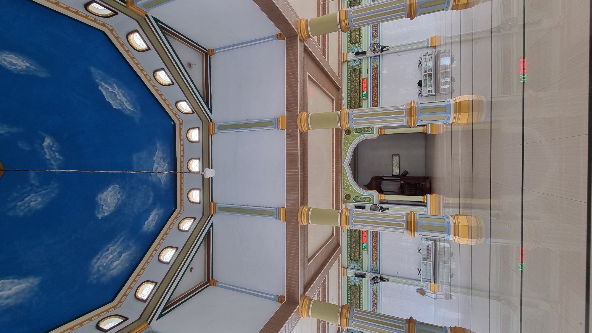 The photograph is a low-angle interior shot of a mosque in Meunasahlhok, Indonesia. The ceiling is painted with a vibrant blue sky and white clouds, with a single light fixture hanging from a cord in the center. The walls are white with decorative borders in green, brown, and gold. Large, fluted columns, painted in alternating sections of yellow, green, and light blue, support the ceiling.
In the middle ground, an arched entrance leads to another part of the mosque. To the left and right of the entrance, Islamic calligraphy is displayed on the walls above digital clocks showing times in red and green. There are also black wall-mounted fans on either side of the entrance. Display cases with books are visible in the background. The floor is made of large, glossy beige tiles that reflect the architecture. The scene is empty of people.