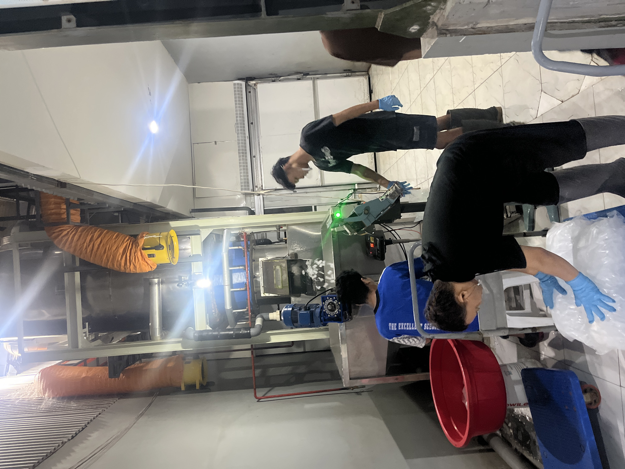 In an indoor industrial setting, possibly a food processing facility in Pekalongan, Indonesia, three individuals are engaged in the operation of an ice-making machine. The machine, a prominent vertical unit with a blue top section, is actively producing ice, which is visible falling into a stainless steel collection area. Orange flexible ducts are attached to the machine, suggesting ventilation or an exhaust system.
Two men, wearing blue gloves, are positioned near the ice output. One, in a blue t-shirt, is observing the process. The other, in a black t-shirt, is interacting with the machine, wearing blue gloves and dark shorts, and tall rubber boots. A third individual, also wearing blue gloves and a black shirt and pants, is focused on bagging the ice into large plastic bags, using a blue dolly and a large red tub nearby for potential ice storage.
The environment appears functional, with tiled flooring, a white wall on the left, and a large white refrigerator unit visible in the background. The lighting is overhead and appears to be a mix of artificial light sources, creating some glare. The overall impression is one of active work in a commercial ice production environment.
