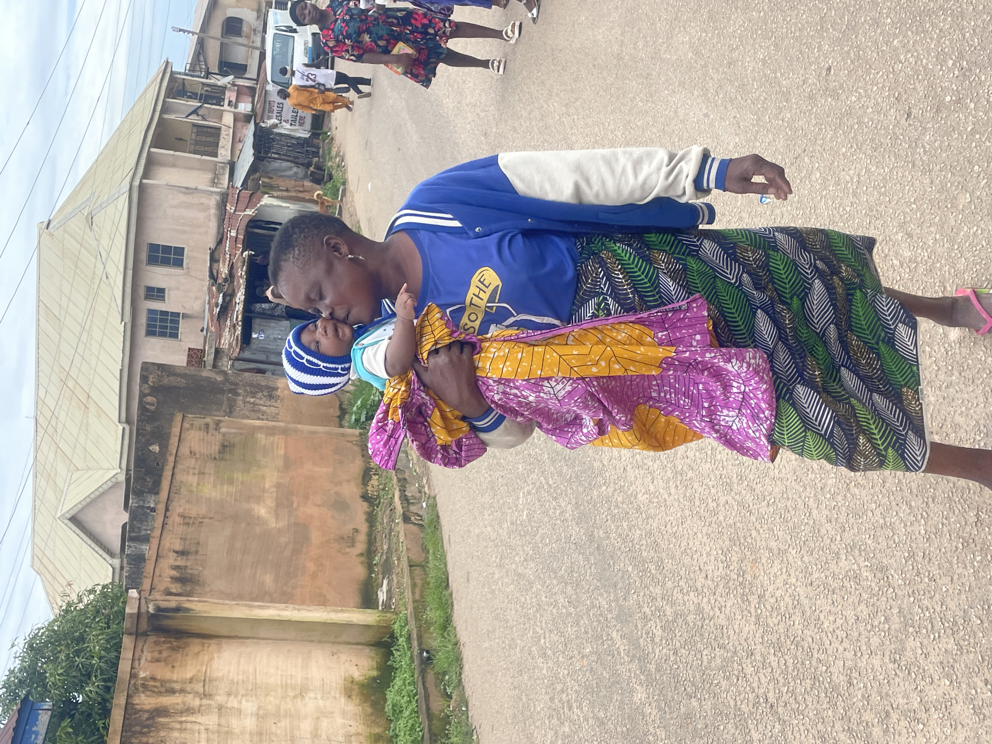 In an outdoor, urban setting in Jos, Nigeria, a woman carries a baby wrapped in a vibrantly patterned fabric. The baby wears a blue and white striped hat and is being kissed by the woman. The woman is dressed in a blue jacket with white sleeves and a green, black, and white patterned skirt. She is wearing pink flip-flops. In the background, buildings line a street, and several other individuals are visible, including a person in an orange outfit and a woman in a floral dress. A white van is also present in the scene. The ground is paved with asphalt.
