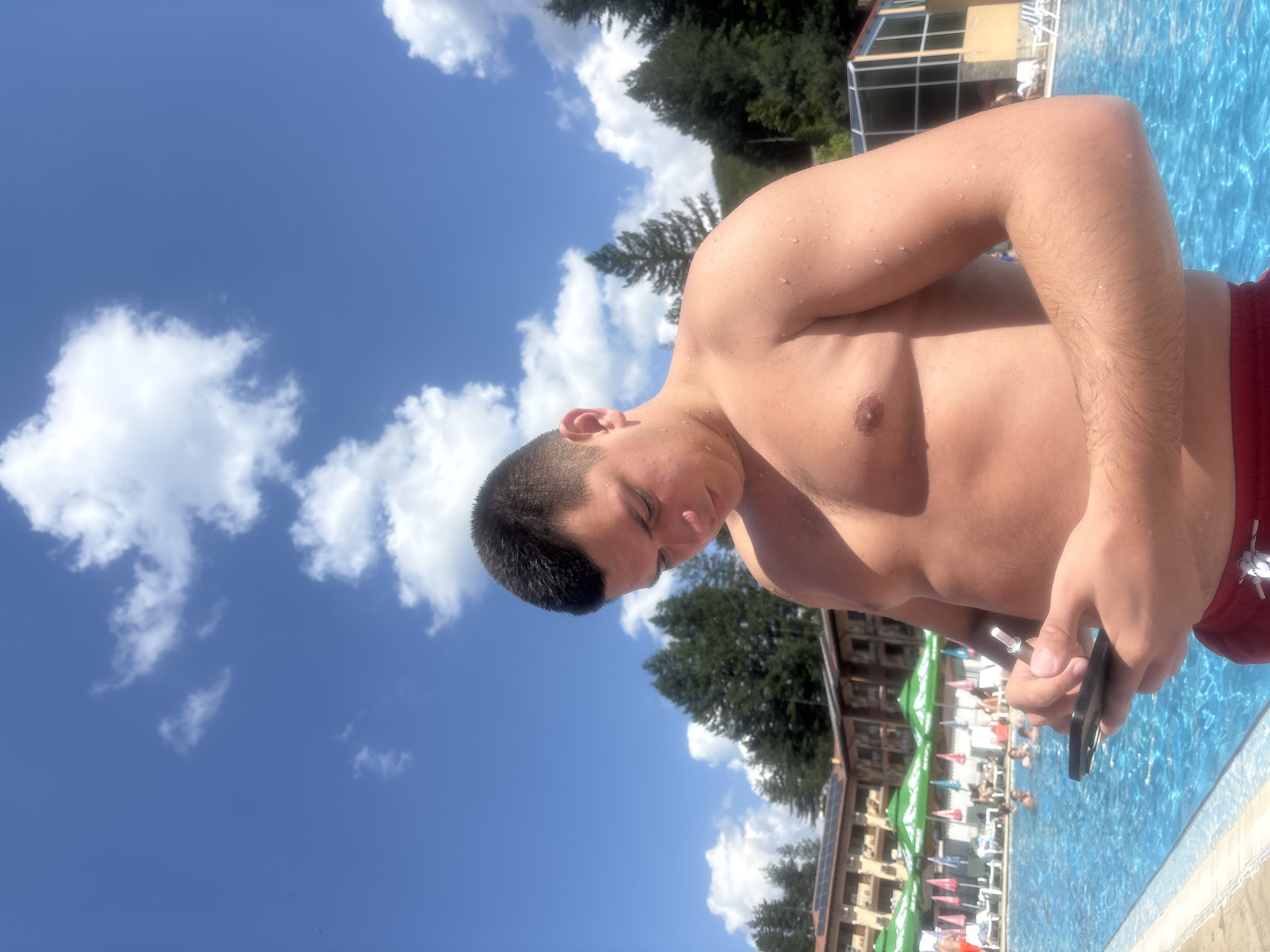 A young man with short, dark hair and a muscular build stands by a bright blue swimming pool, looking down at his phone. Water droplets glisten on his bare skin. He is wearing red swim trunks. In the background, a resort building with a red-tiled roof and solar panels is visible, along with rows of green and pink umbrellas providing shade over lounge chairs. Several people are in the pool and relaxing by the poolside. Lush green trees surround the resort, and a clear blue sky with fluffy white clouds stretches overhead. The scene is set outdoors during the day in Chiflik, Bulgaria.
