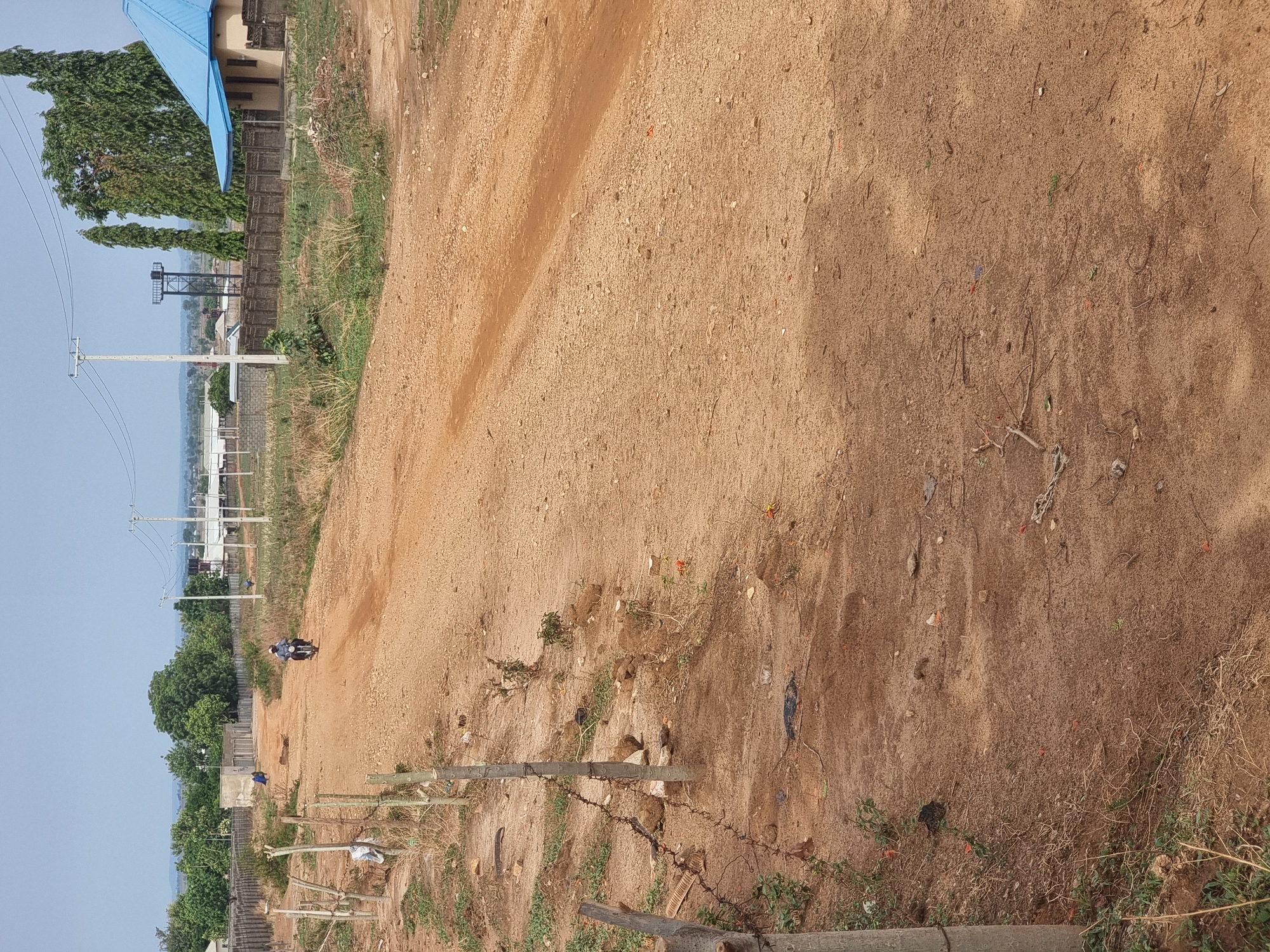 The image depicts a dirt road in Gwagwalada, Nigeria. A motorcycle is travelling on the road. The environment includes sparse vegetation, a fence with barbed wire, and electrical poles in the distance. A building with a blue roof is visible in the background. The sky is light blue.