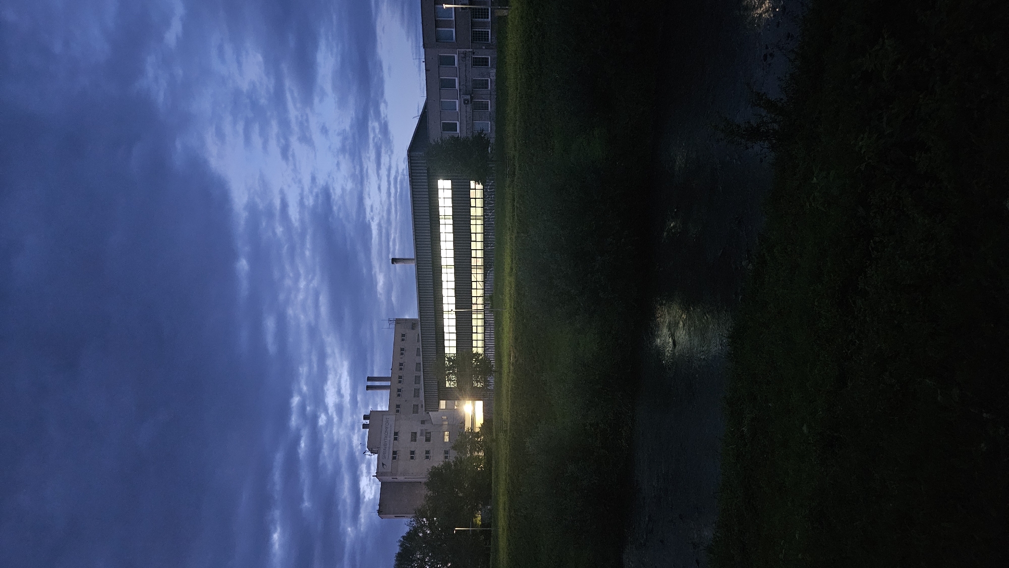 The image depicts an industrial building at dusk in Prešov, Slovakia. The building features multiple rows of illuminated windows, suggesting interior lighting. There are smokestacks visible on top of one section of the building, indicating industrial activity. In the foreground, a body of water, possibly a river or canal, reflects the dim light from the sky and the building. The surrounding vegetation is dark, and the sky is filled with dramatic, dark clouds, typical of a stormy evening. The scene is devoid of any visible human presence. The building's facade displays some text, which appears to read "SPRAYBYTKOMFORT" on the upper section.