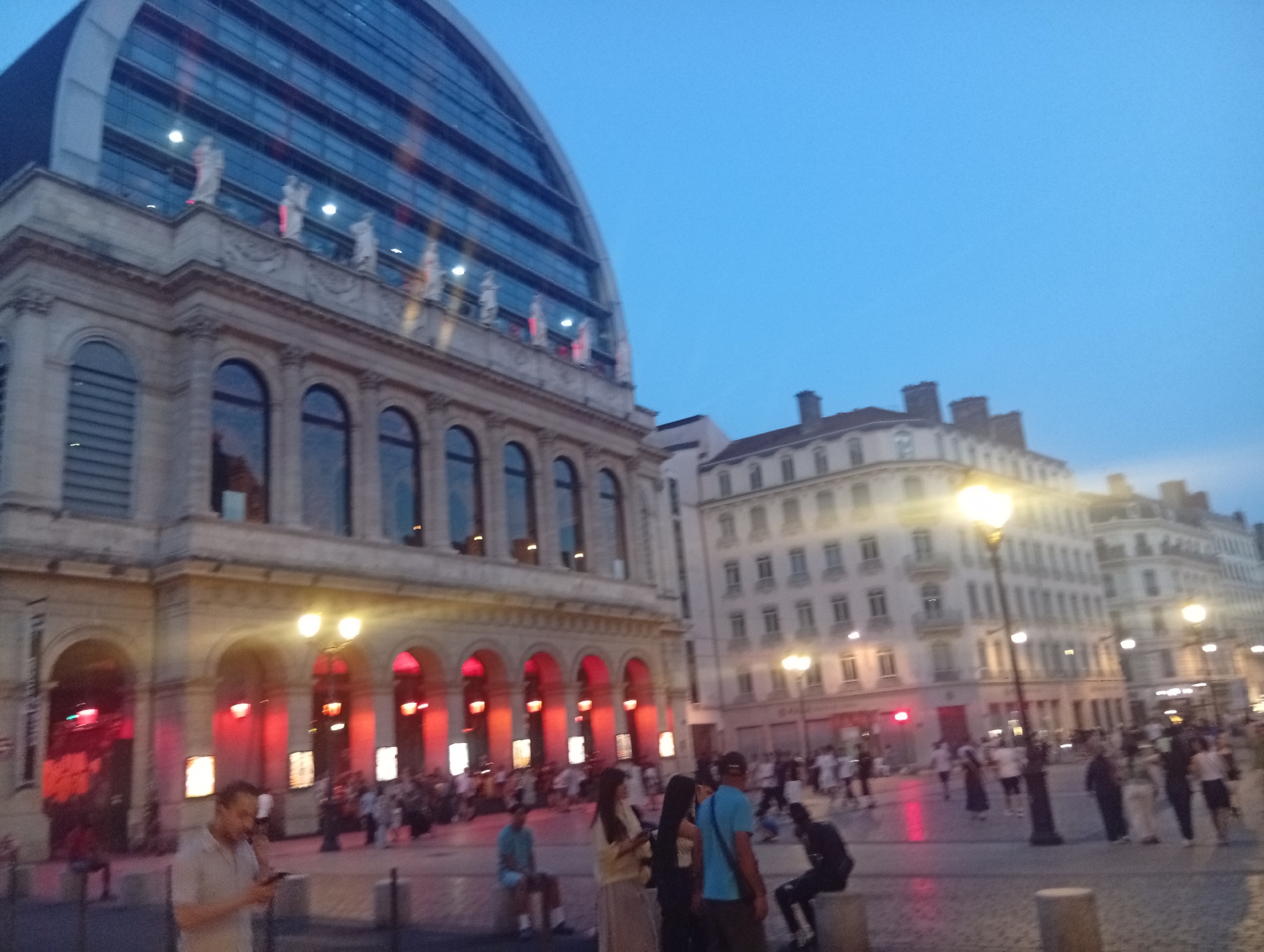 The image depicts Place Bellecour in Lyon, France, at dusk. The iconic opera house with its modern glass facade is visible on the left. The main building features classical architecture with arched windows and decorative statues. In the foreground, a man in a light-colored shirt is looking at his phone. The square is bustling with people walking and gathering. Several streetlights illuminate the scene, casting a warm glow on the pavement and buildings. The sky is a clear, pale blue, indicating the transition from day to night. The overall atmosphere is lively and urban.