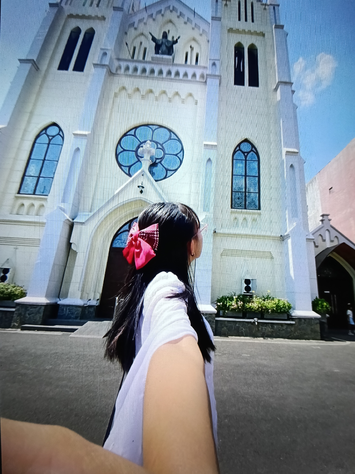 The image shows a woman with long dark hair and a pink bow on her head, facing away from the camera and towards a large, white church. The church has a tall, ornate facade with arched windows and a circular stained-glass window above the entrance. The woman is wearing a white shirt and appears to be holding the camera out in front of her, taking a selfie. In the background, there is a blue sky with a few clouds. The church is identified as the Gereja Katedral Santa Maria Diangkat Ke Surga, located in Raha, Indonesia. The surrounding area appears to be an urban environment with a paved walkway and some greenery.