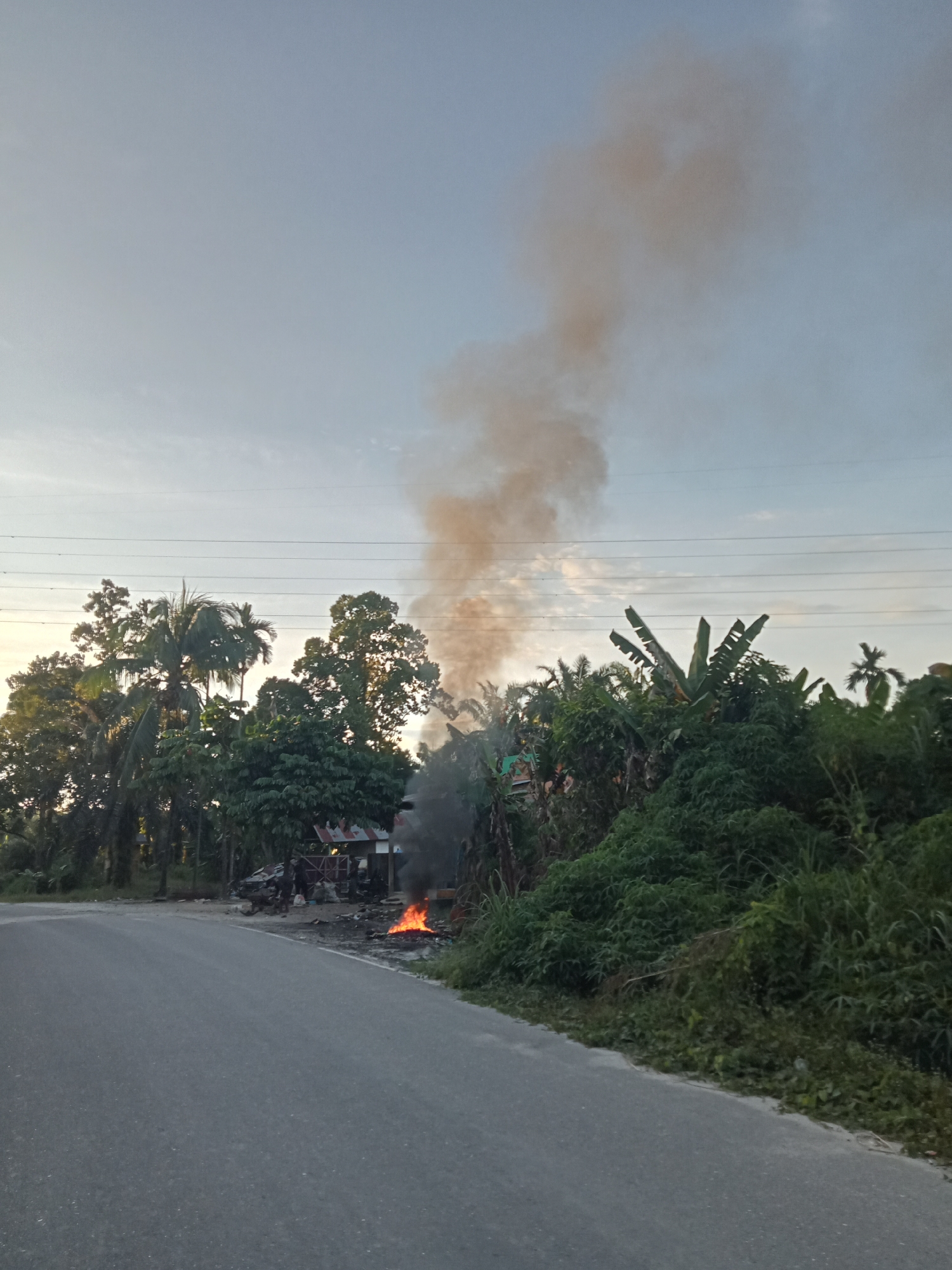 In Kualu, Indonesia, by the side of a paved road, a fire burns. The fire is located near a small structure with a red-and-white corrugated metal roof, surrounded by debris. A plume of smoke rises from the fire, drifting upwards into the sky. Several trees, including palm trees, are visible in the background, and lush green vegetation lines the right side of the road. Overhead, power lines are strung across the sky. The scene appears to be at dusk or dawn, with a pale sky.