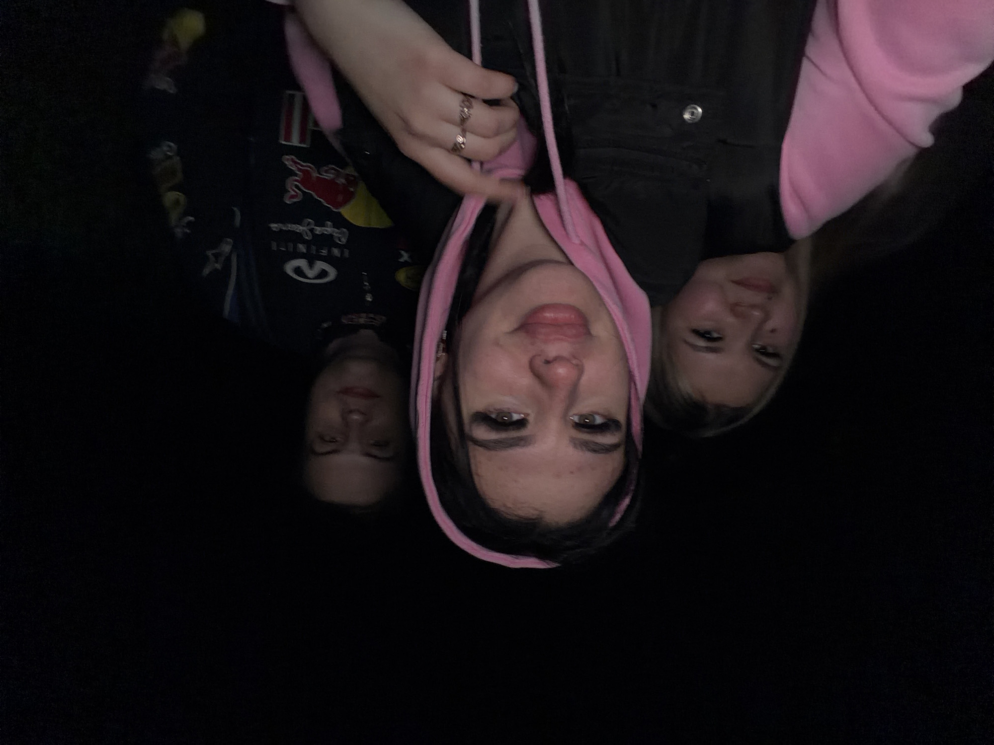 Three women are pictured in dim lighting. The woman in the center wears a black vest over a pink hooded sweatshirt. She is pulling the hood up over her head. A hand is visible near the hood, with a ring on the index finger. The woman on the left wears a dark long-sleeved shirt with racing-themed logos. The woman on the right has blonde hair and wears a pink sweatshirt. The background is dark and indistinct. The image's orientation suggests the women are holding the camera above them and pointing it downwards, but the specific location is not determinable from the image.