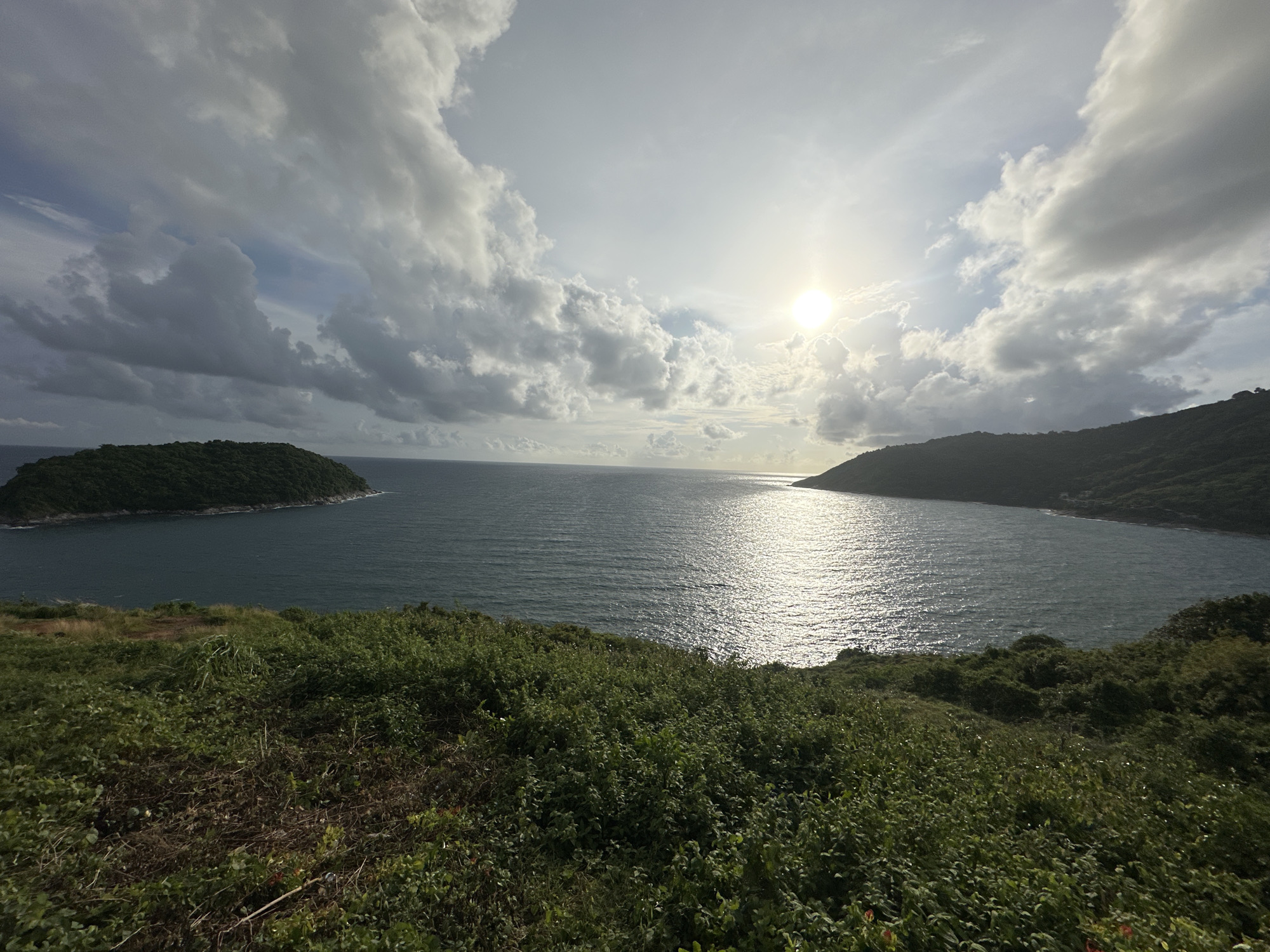 The image depicts a scenic view of the ocean and coastline, likely taken from an elevated vantage point. In the foreground, there is lush green vegetation covering a sloping hill. The middle ground features a body of calm, dark blue water, reflecting the sunlight. On either side of the water, hills covered in dense green foliage rise from the sea, forming a bay or cove. The sky above is a mix of bright blue and dramatic grey clouds, with the sun shining brightly, casting a shimmering reflection on the water. The overall impression is one of natural beauty and tranquility. The location is identified as Rawai, Thailand, suggesting a tropical coastal environment. There are no people or man-made structures clearly visible in the image, emphasizing the natural landscape.