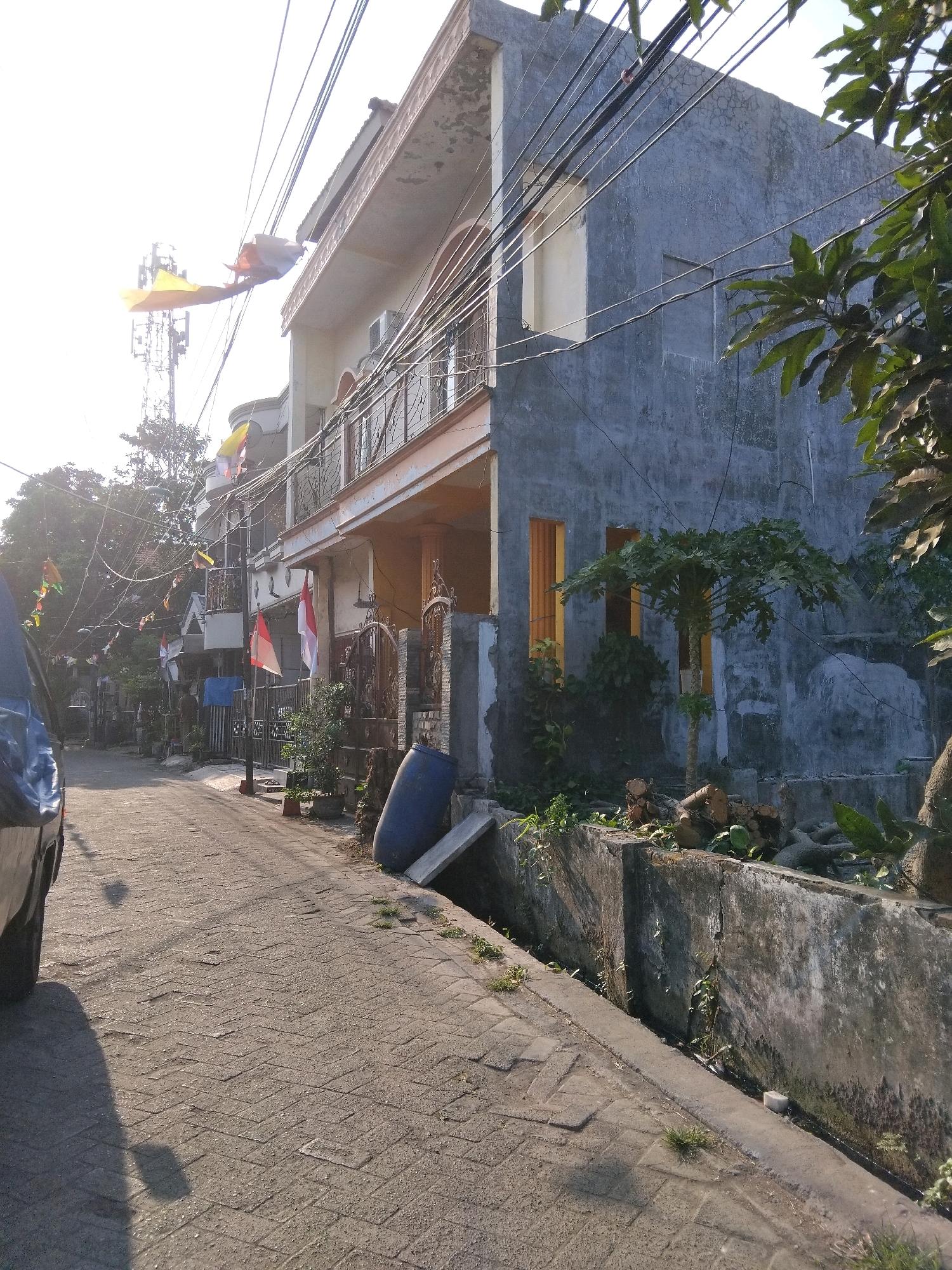 A street scene in Berbek, Indonesia, features a paved road lined with residential buildings and vegetation. The image captures a two-story house with a textured gray exterior, a light-colored trim, and visible electrical wires overhead. In front of the house, a balcony with metal railings overlooks the street. Various flags, including Indonesian flags, are displayed along the street and on the buildings, suggesting a celebratory occasion. The road is constructed with interlocking pavers, and a gutter runs along the right side of the road. A large blue barrel and some cut logs are present near the gutter. Trees and foliage are visible throughout the scene, with a telecommunications tower in the background. The lighting suggests it is either early morning or late afternoon, with long shadows cast across the road. A dark-colored vehicle is partially visible on the left side of the image.
