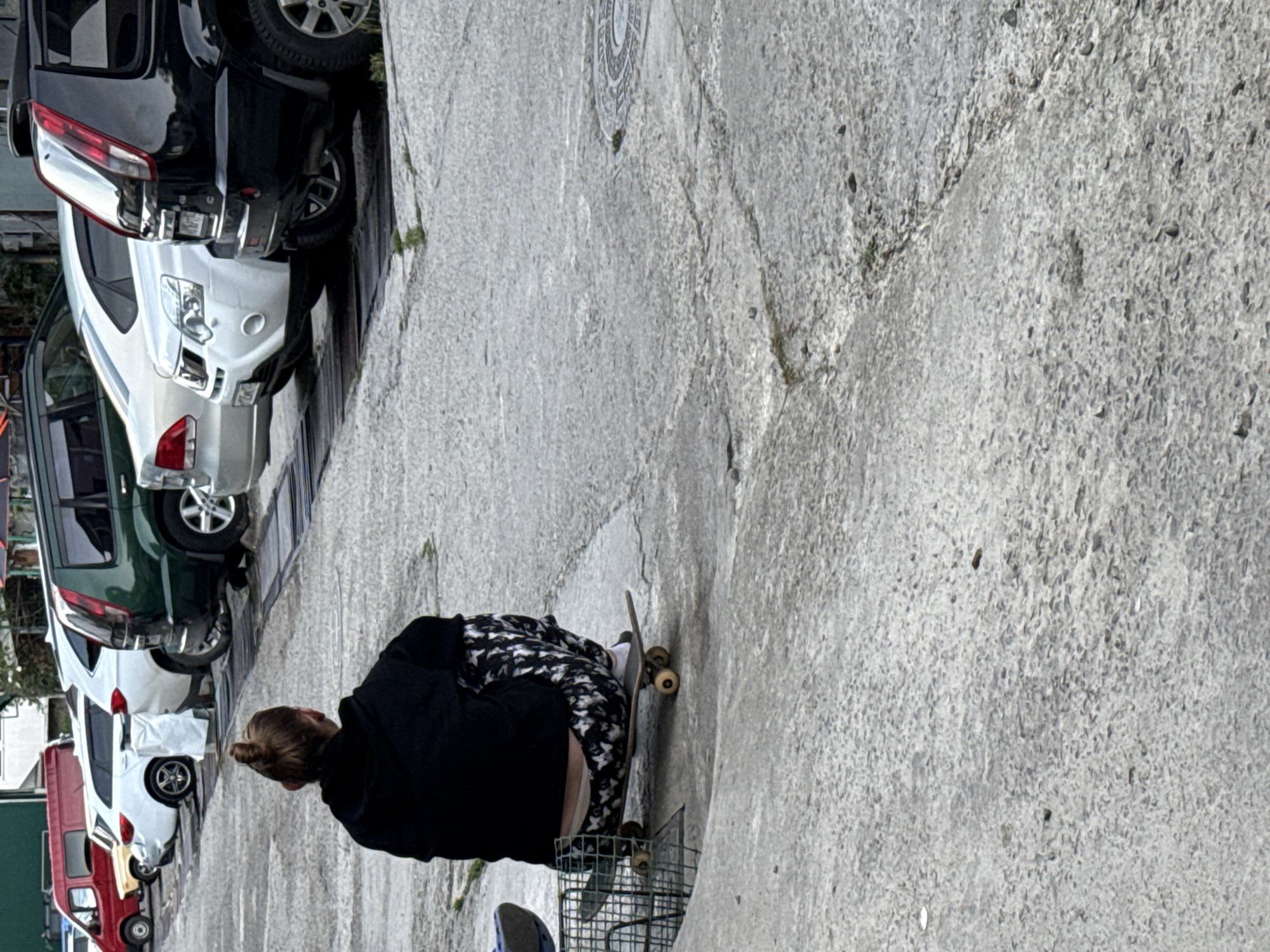 A person crouches on a skateboard in the city of Unknown, Unknown, next to a wire cage and a row of parked vehicles. The person wears a black sweatshirt, printed pants, and white socks. The skateboard is resting on a concrete surface. Parked cars line the left side of the frame. The environment seems to be an urban street or parking area.