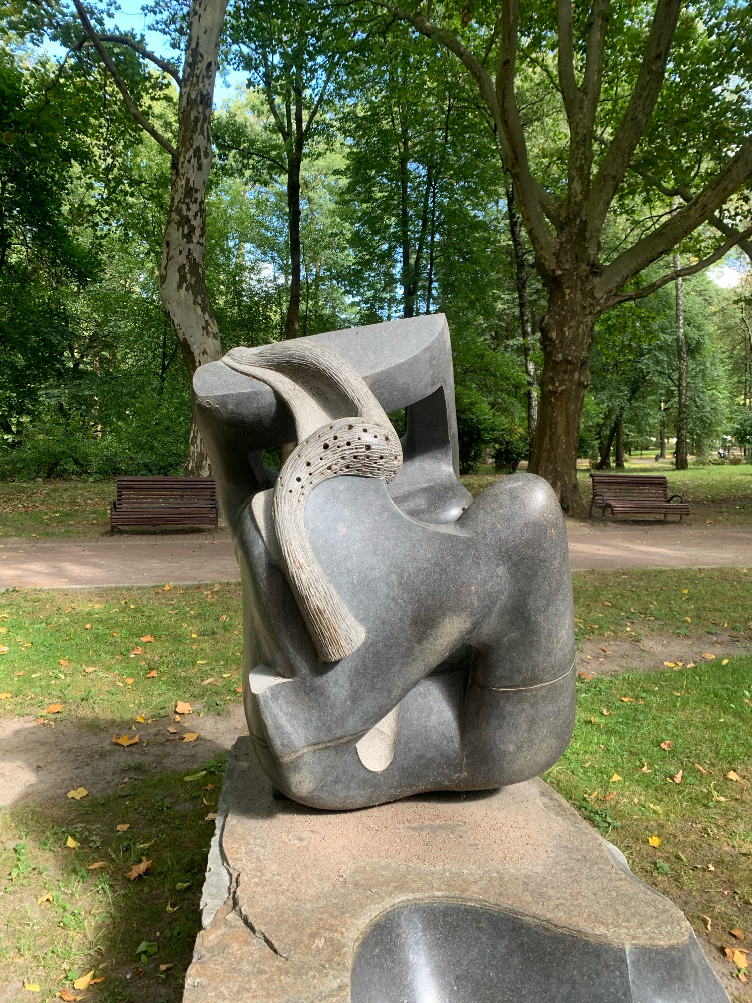 The image depicts a modern abstract sculpture located outdoors in a park setting. The sculpture, made of dark, polished stone, has a rounded, organic form that suggests a seated or reclining figure. A textured element, resembling a rope or fabric, is draped over a section of the sculpture. The sculpture rests on a rough, irregular stone base.
The park environment features lush green grass, scattered fallen leaves, and a paved pathway. In the background, several large trees with textured bark and dense green foliage dominate the scene. Two wooden park benches are visible in the mid-ground. The sky is partially visible through the tree canopy, appearing bright and possibly sunny.
This sculpture is located in Lviv, Ukraine, specifically in the Ivan Franko Park.