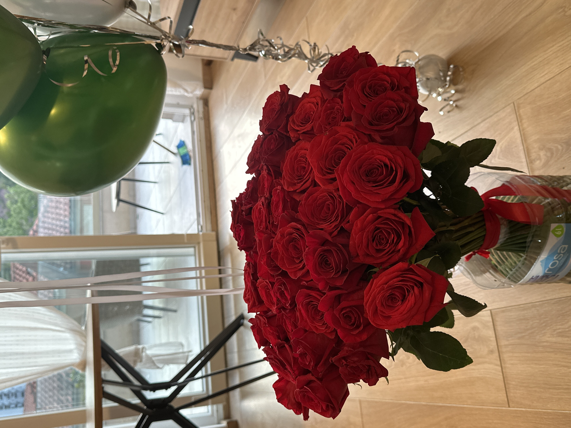 The image shows a bouquet of red roses in a clear plastic container with a red ribbon tied around it, sitting on a light-colored wooden floor. To the left of the bouquet are green and white balloons tied with silver ribbon. In the background, a window with white frames and blinds is visible, offering a glimpse of a cityscape. The location is Donji Pobori, Montenegro, as inferred from the setting visible through the window. A white laundry drying rack is placed next to the window.