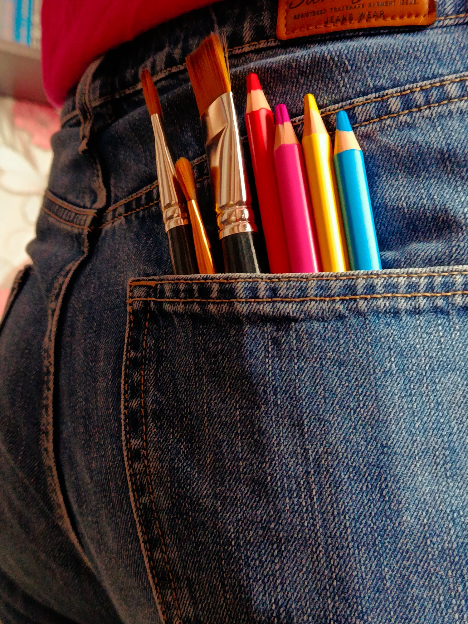 A close-up shot shows a pair of blue jeans with various art supplies sticking out of the back pocket. These include three paintbrushes with black handles and gold ferrules, and four colored pencils in the following colors: red, pink, yellow, and blue. A leather patch with text on it is visible above the pocket. The background is slightly blurred. The city of Фоки, Russia is not relevant or present in the image's content.