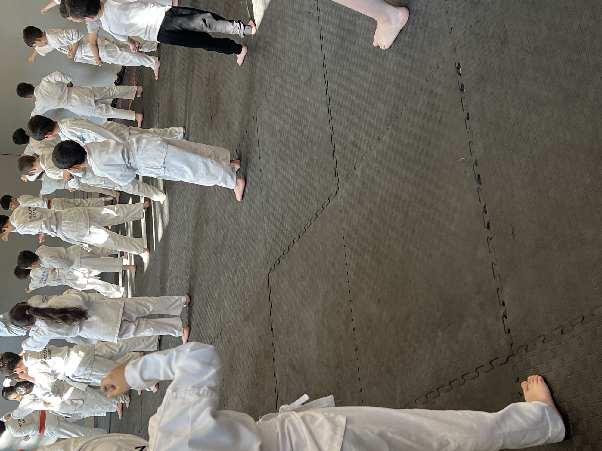 The image depicts a group of children and adolescents practicing martial arts, specifically karate, in an indoor training facility. The participants are wearing white karate gi uniforms and are barefoot, standing on a black, interlocking puzzle mat floor. The room appears to be a gymnasium or dojo, with a plain gray wall in the background. Sunlight streams in from an unseen source, casting shadows and illuminating the scene. The children are arranged in rows, some facing forward and others turned slightly, suggesting they are in the middle of a lesson or demonstration. Their stances and arm positions indicate active participation in training. One child in the foreground is partially visible, their white uniform and bare feet prominent. Another child to their right is also in focus, wearing a white gi and black pants, indicating a possible difference in skill level or uniform style. The overall atmosphere is one of disciplined activity and learning. The location is identified as a karate dojo in Vanadzor, Armenia.