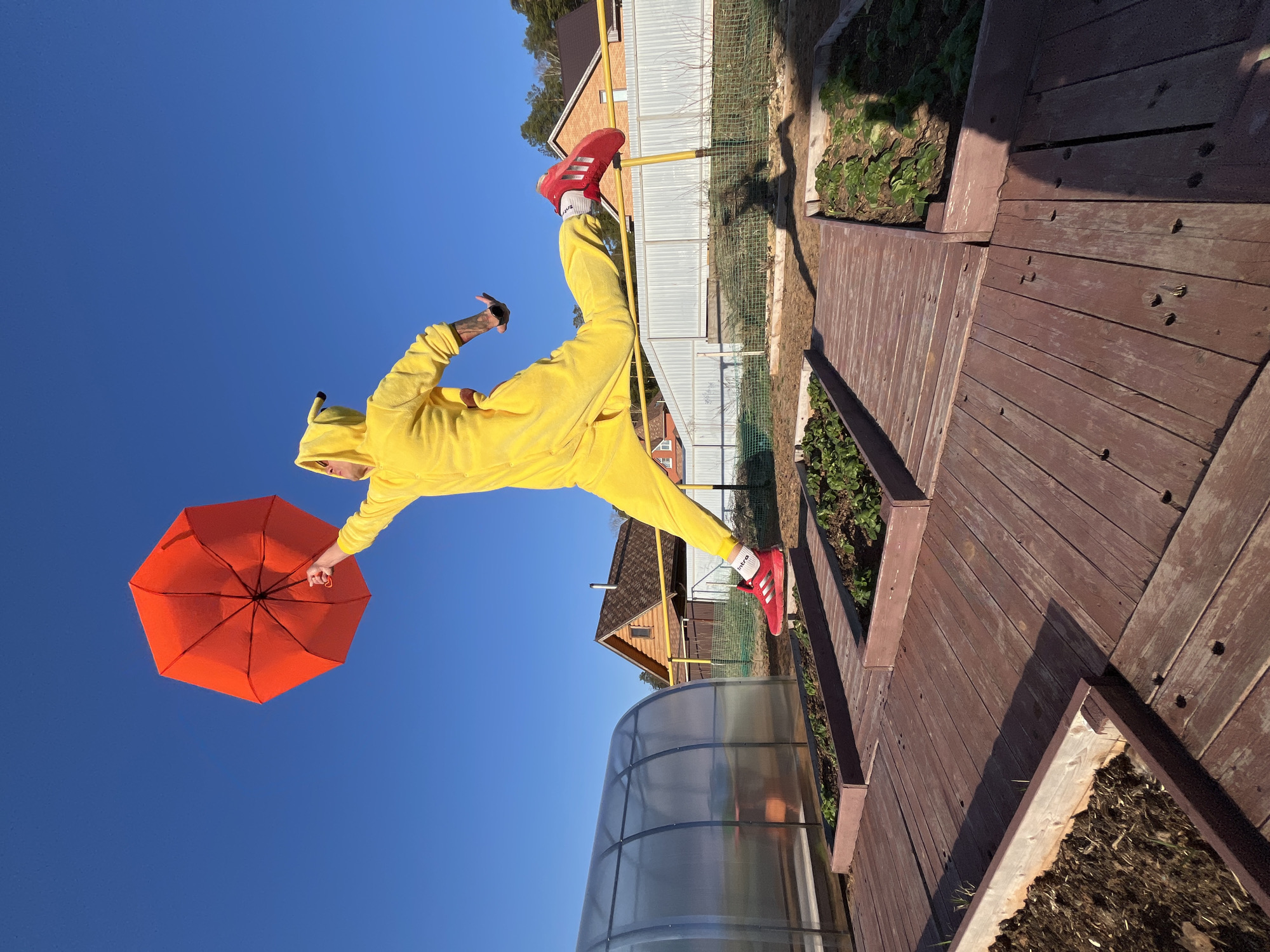 A person wearing a yellow Pikachu onesie and red sneakers is suspended in mid-air, seemingly balancing horizontally in a photograph. They are holding an orange umbrella. Behind them, there's a clear blue sky. In the background are elements of a domestic setting including: a wooden walkway, garden beds, a greenhouse, a metal fence, houses with pitched roofs, and trees. The location appears to be the city of Chaikovsky, Russia, based on the presence of architectural elements and plants.