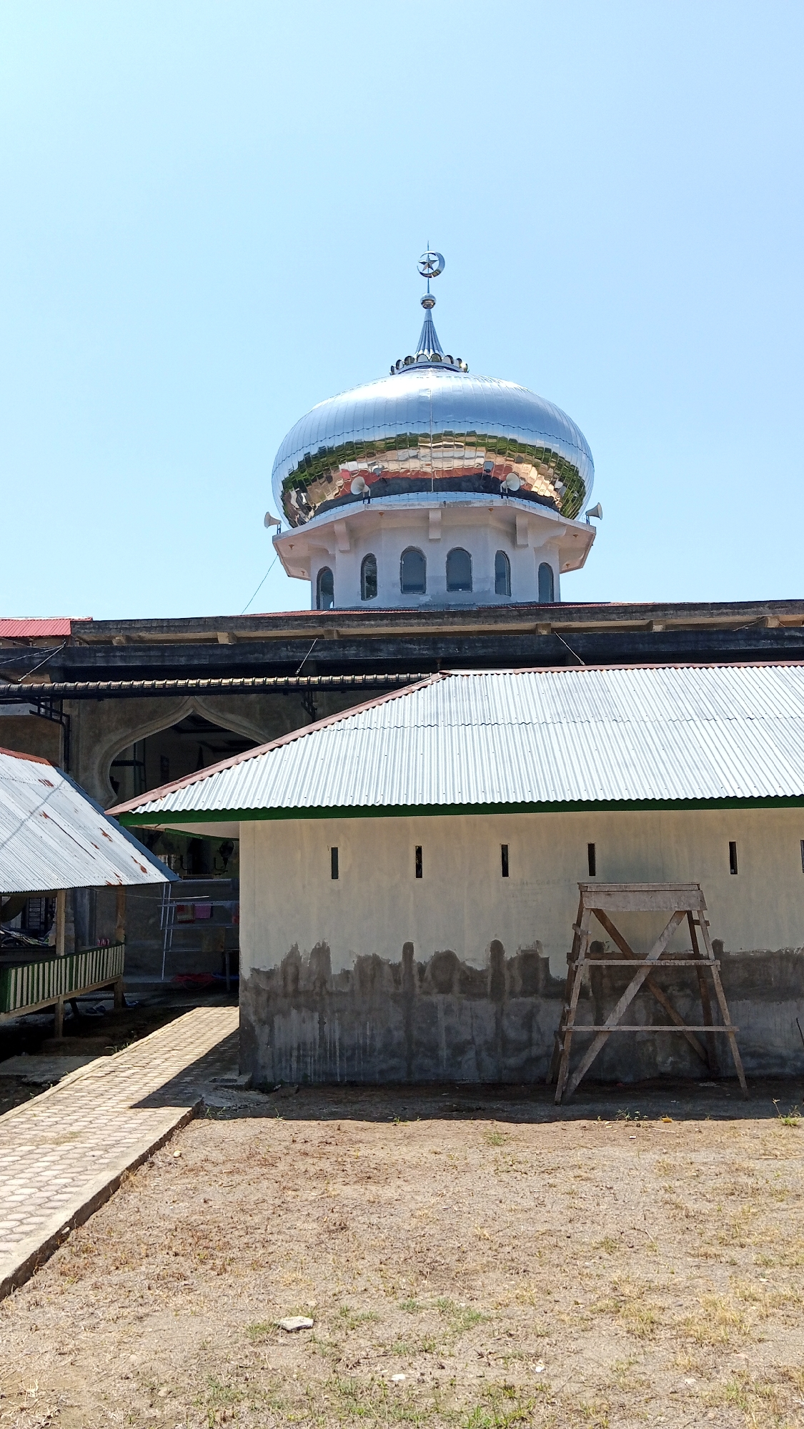 The image shows a mosque with a prominent, silver, dome-shaped roof topped with a crescent moon and star. The dome reflects the surrounding environment, including buildings and trees. In the foreground, there is a building with a corrugated metal roof, a light-colored wall with a rough, grey, lower section, and a wooden A-frame ladder leaning against it. To the left, a paved pathway leads towards another structure with a similar corrugated metal roof. The ground is dry and covered in sparse, brown vegetation. The sky is clear and blue. The scene appears to be in Uteunreutoh, Indonesia.