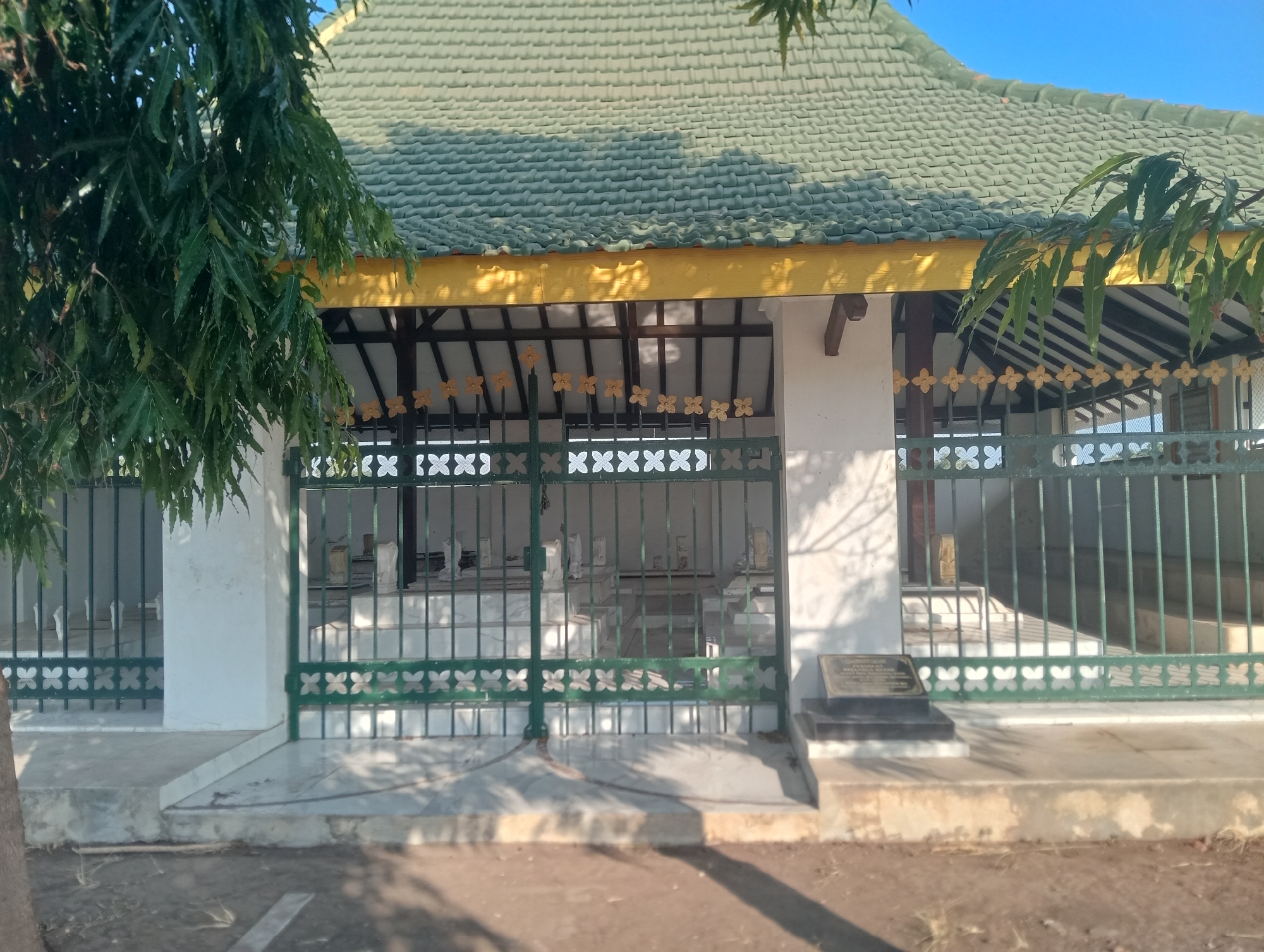 The image shows a well-maintained grave complex located outdoors, possibly a cemetery or a tomb of historical significance, in Bloro, Indonesia. The structure features a green tiled roof with a yellow trim, supported by dark wooden beams. A green metal fence with a decorative pattern of four-leaf clovers encloses the area where several white tombstones are visible. Some of the tombstones are upright, while others appear to be flat slabs. A small, dark plaque is mounted on a granite base outside the fence, suggesting it might commemorate the person or people buried within. The grounds are paved with white marble or similar material, and the surrounding area appears to be dirt or gravel. A large tree with green, feathery leaves partially obscures the left side of the image. The sky is clear and blue, indicating fair weather. The scene is illuminated by sunlight, casting shadows on the ground and the structure.