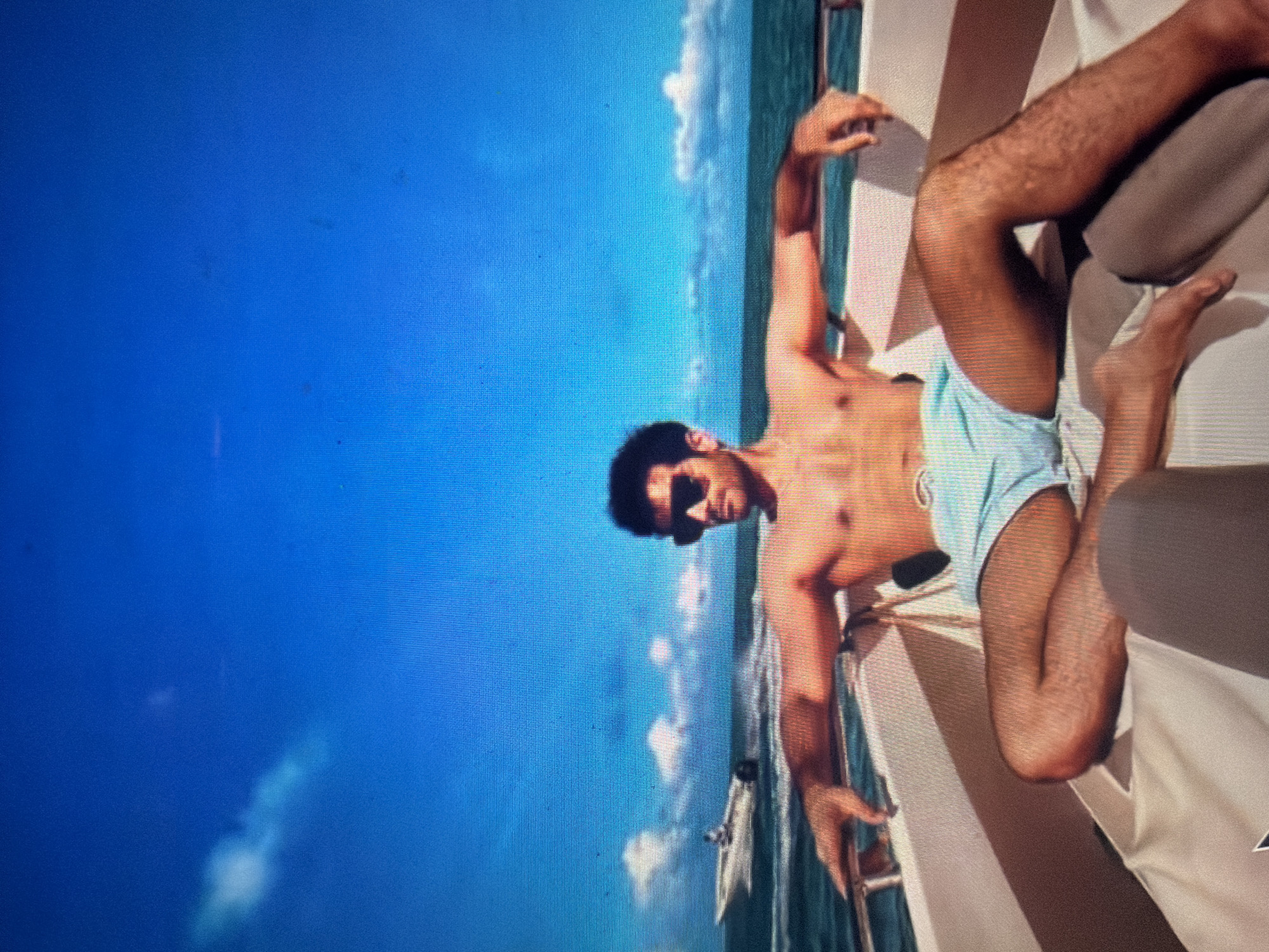 The image depicts a scene located in the city of Shumen, Bulgaria, featuring a shirtless male individual relaxing on a white motorboat. The subject, positioned centrally in the foreground, is a light-skinned male with dark, curly hair and dark rectangular sunglasses. He wears light blue swim shorts. His arms are spread wide, with his forearms resting on the white gunwales of the boat. His legs are bent and splayed, with bare feet visible. The boat's interior is upholstered in a light beige material. Behind the subject, a vast expanse of deep blue water with light choppiness is visible, extending to the horizon. In the mid-ground, to the left, another white powerboat is seen moving away, leaving a white wake behind it. The sky above is a vibrant, clear blue with a few scattered white cumulus clouds, particularly on the left side. The entire image exhibits visible screen pixelation and minor dust or defect artifacts, indicative of a photograph of a digital screen.