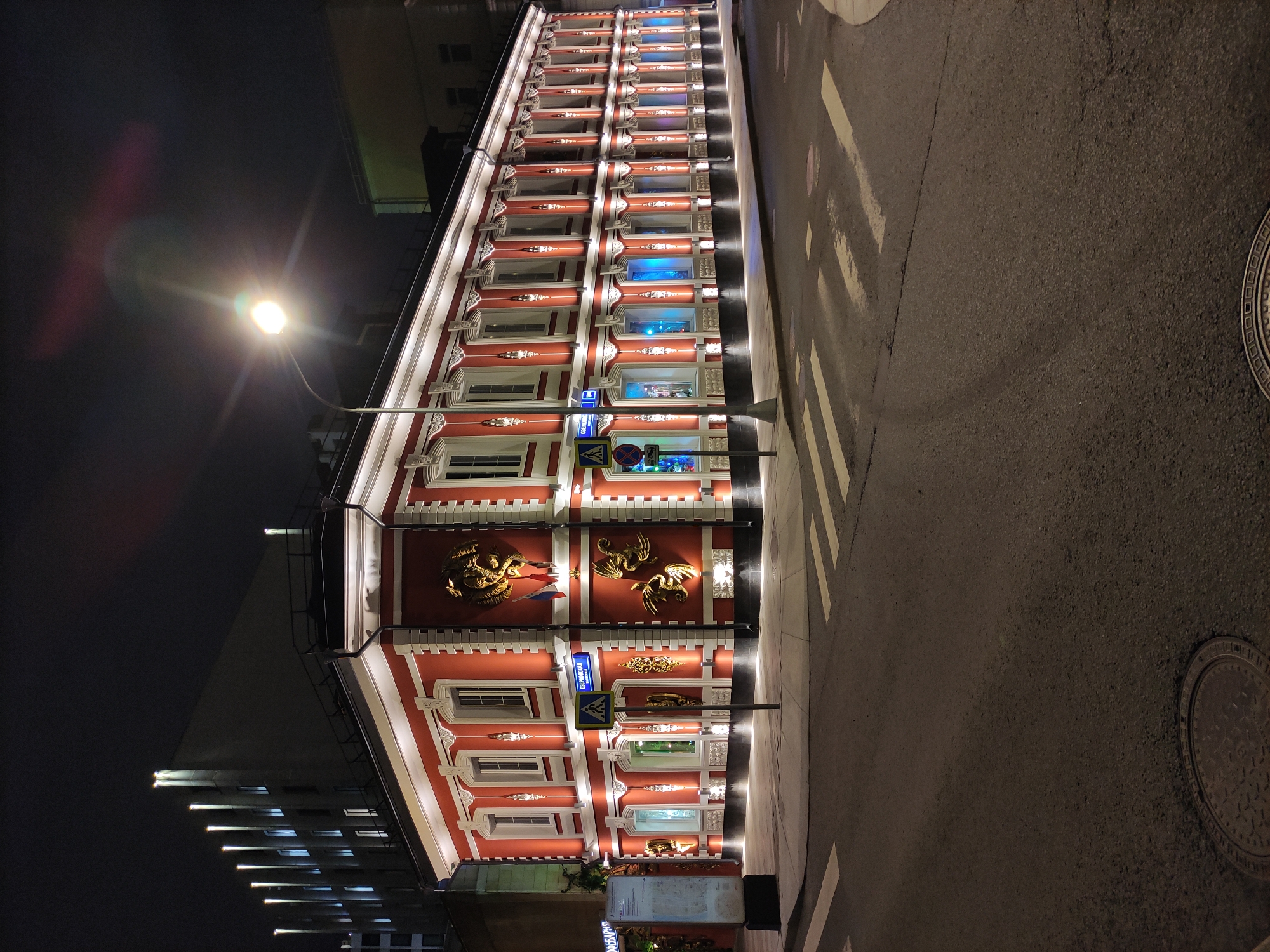 The image shows a building at night. The building is red and white and has multiple windows. There are some golden decorations on the facade. A streetlight is in the upper-left part of the image. To the right of the building is a paved road with white stripes. A manhole cover is in the road.