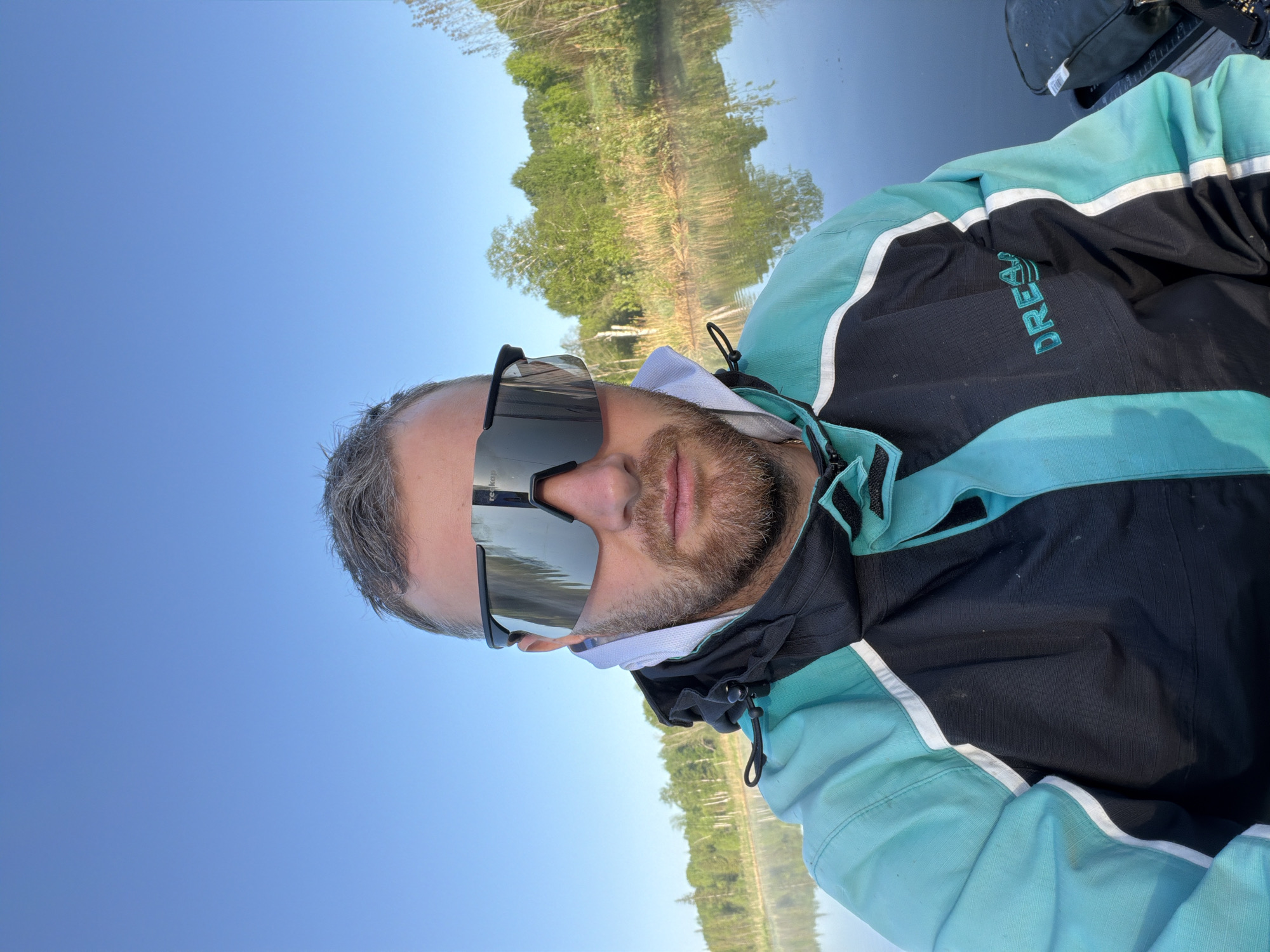A man with graying hair and a beard wears dark sunglasses and a black, white, and light blue jacket with the word "DREAM" embroidered on it. He appears to be near a body of water, possibly a lake or river, with trees visible in the background and their reflections mirrored in the water. The sky is clear and blue. The image appears to be taken in the daytime. The location is not identifiable as Belaya Gora, Russia, based on the given visual information.
