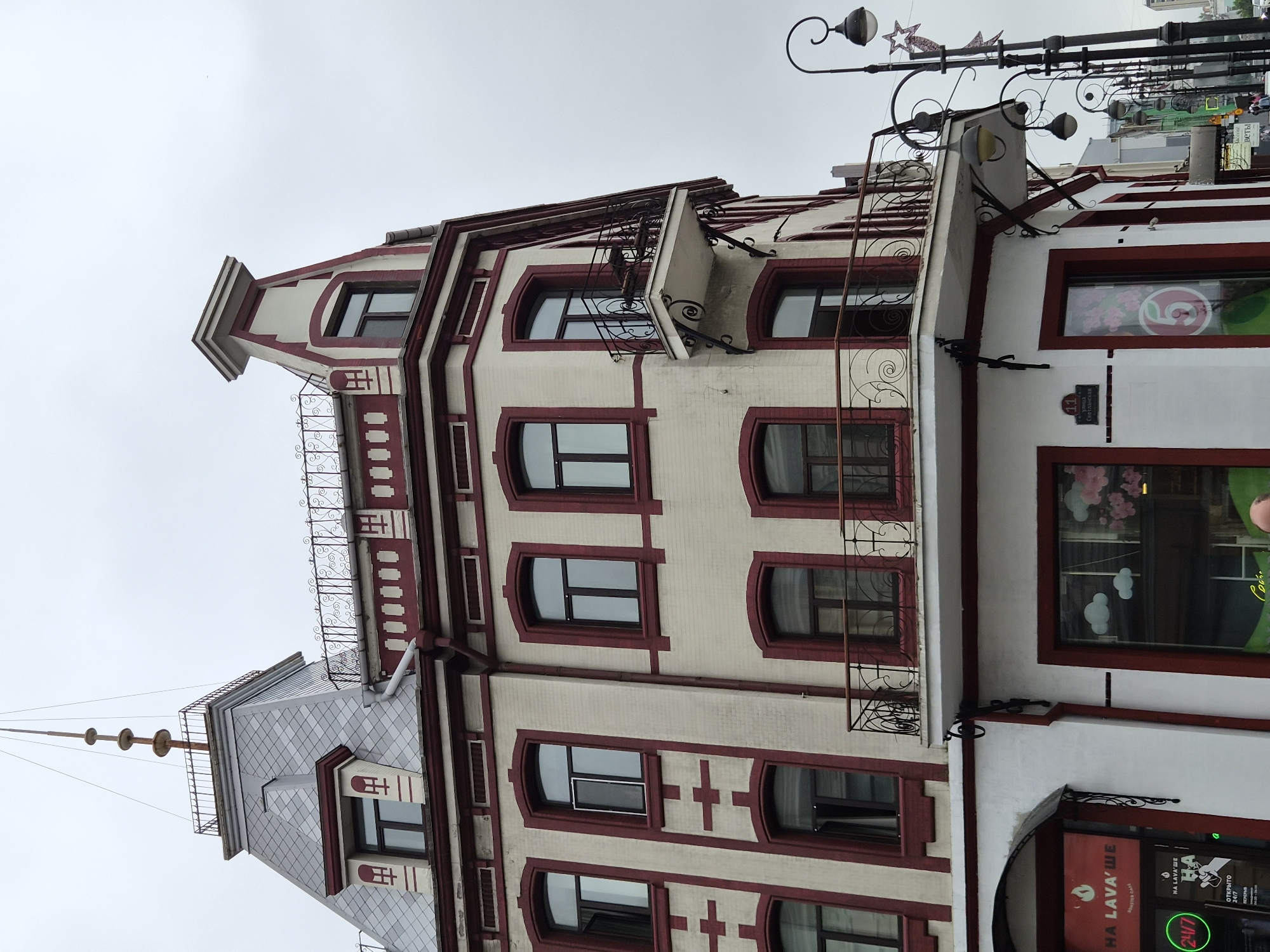 The image depicts the facade of a multi-story building with a distinctive corner design, featuring a pointed turret topped with a spire and intricate metalwork. The building's exterior is cream-colored with deep red trim around windows, balconies, and decorative elements. Several windows are visible, some with dark frames, and a few are open. Two balconies with ornate wrought-iron railings are present, one on the corner of the building and another extending from the side. The lower level of the building appears to house commercial establishments, with a prominent sign that reads "НА LAVA'ШЕ ROASTER CAFE" and a "24/7" indicator. Streetlights with ornate curves and globe-shaped fixtures line the street in front of the building. The sky is overcast, suggesting a cloudy day. In the background, other buildings and street elements are visible, including a street sign that says "11 улица Светланская," indicating the location as Svetlanskaya Street, house number 11, in Vladivostok, Russia. A few individuals can be observed in the distance on the sidewalk.