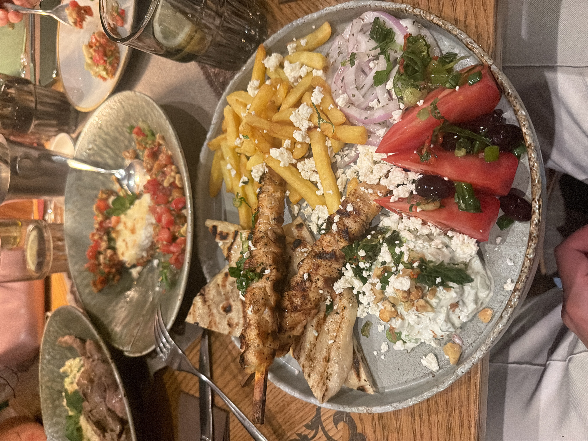 This overhead shot captures a dining scene featuring a variety of Greek-inspired dishes served on a rustic wooden table in Sofia, Bulgaria. The central focus is a large, round grey platter laden with a main course and sides. On this platter, two chicken souvlaki skewers are presented on grilled pita bread. Accompanying these are a generous portion of golden French fries topped with crumbled feta cheese. A fresh salad section includes sliced red onions, wedges of ripe tomatoes, black olives, and fresh herbs. A creamy white dip, likely tzatziki, is also on the platter, garnished with crumbled feta, herbs, and what appear to be crunchy nuts or croutons.
To the left of the main platter, two smaller oval plates are visible. One contains a red and white dish, possibly a tomato salad or dip, with a spoon resting in it. Below that, another plate holds shredded meat with a green accompaniment, possibly a sauce or herb mixture. A person's hand holding a fork is visible at the top left, reaching for food from one of the smaller plates. Another person's arm is partially visible on the right side of the main platter.
A clear glass filled with water and lemon slices is seen in the upper left corner. The overall setting is that of an indoor restaurant or eatery, suggested by the table, plates, and cutlery. The lighting appears warm, indicating an evening meal. There is no visible text in the image.